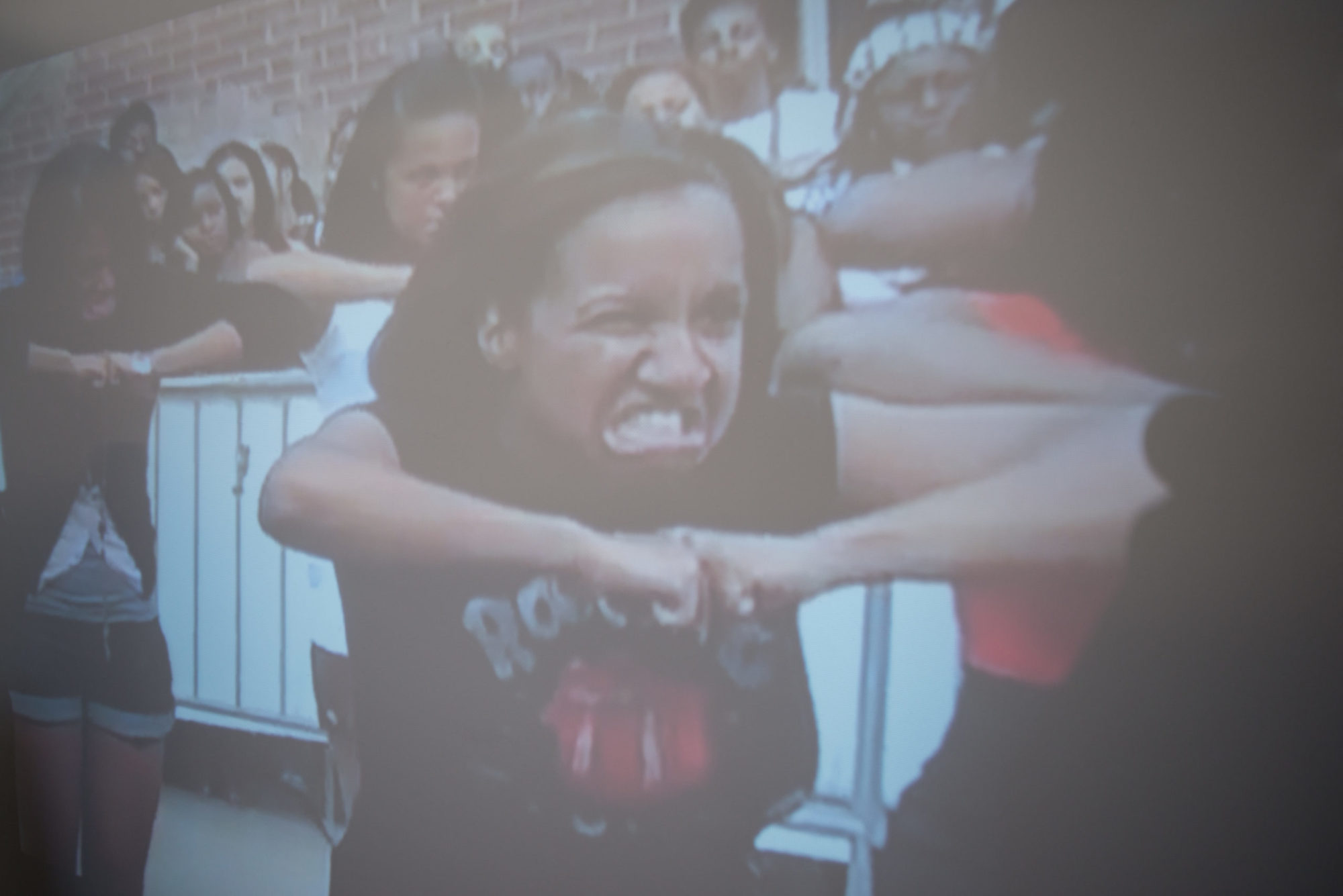 A photo of several people gritting their teeth with elbows raised and fists together. The centermost figure is wearing a black and red T shirt, has shoulder-length hair and seems as if they are in pain.