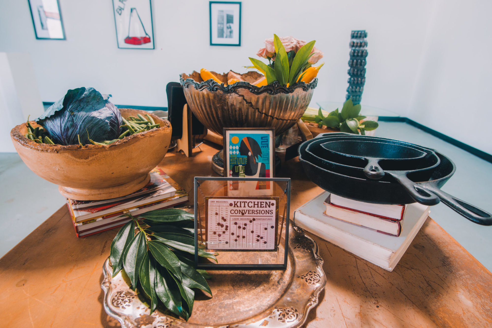 A photo of a wooden table in a room with white walls. Several bowls, plates and pans sit on the table. Some of them are resting on stacks of books. In the bowl closest to the walls there are flowers and fruit. In the plate closest to the viewer there are green leaves and a framed plaque that reads 