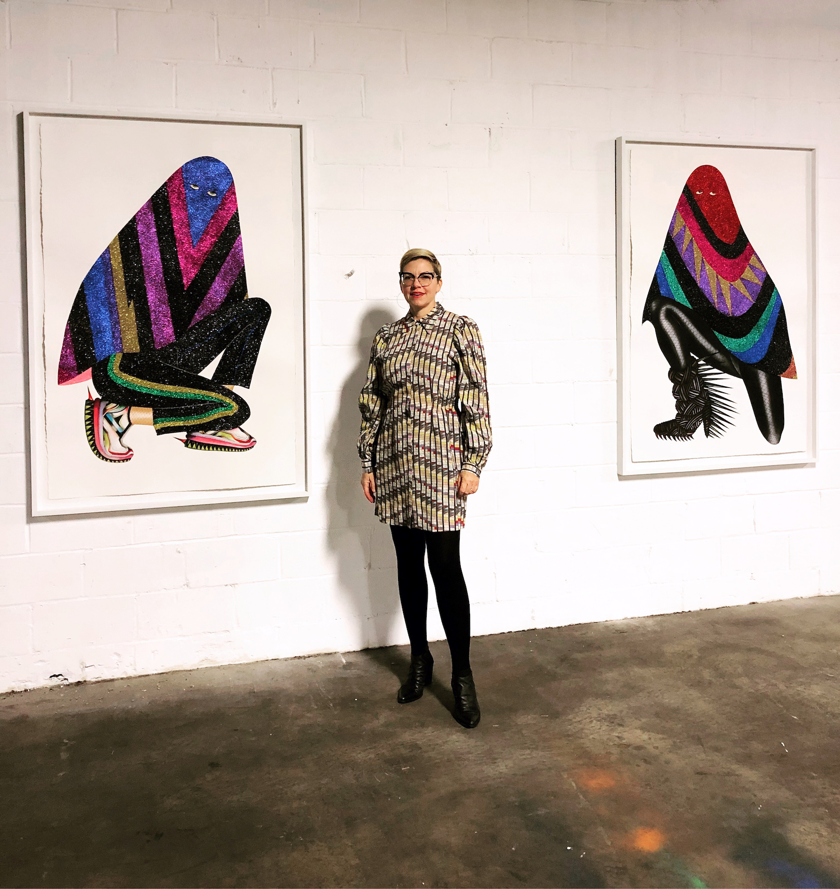 Jen Ray stands in between two of her pieces, each piece depicting a person with their face covered by a cloth.