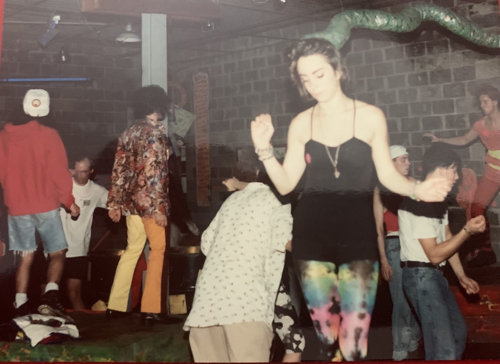 A photograph of individuals in a room with a brick wall. The centermost individual is wearing a black tank top, colorful pants and a fake snake on their head. Nobody is looking at the camera.