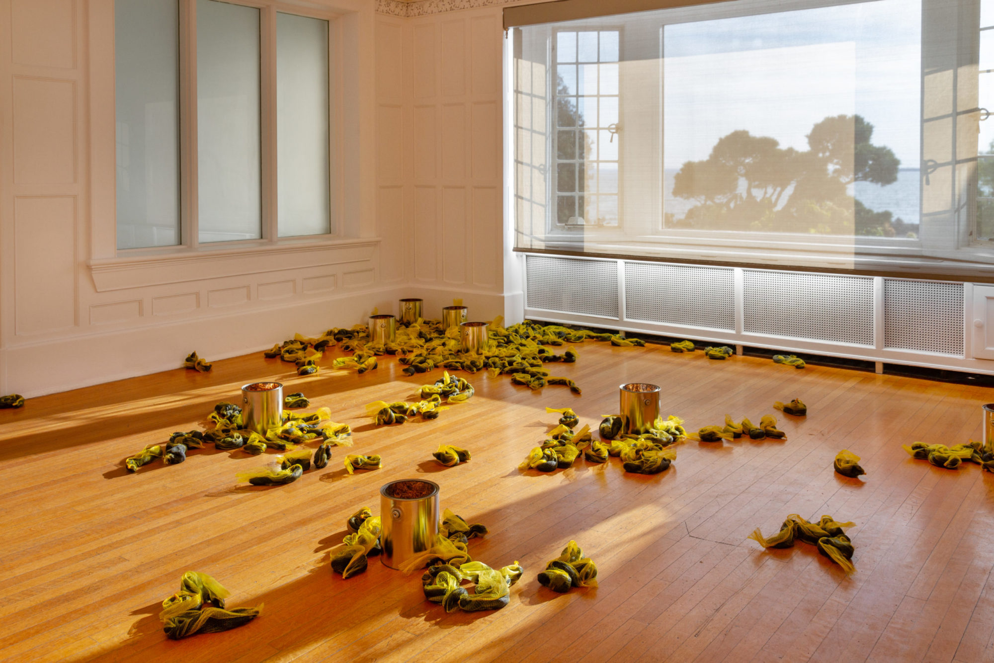 Installation shot of Laurie Kang's work entitled 'Bloom' - black polymer clay 'worms' encased in yellow mesh lay scattered around a room - the clay pieces centralize around silver reflective paint cans