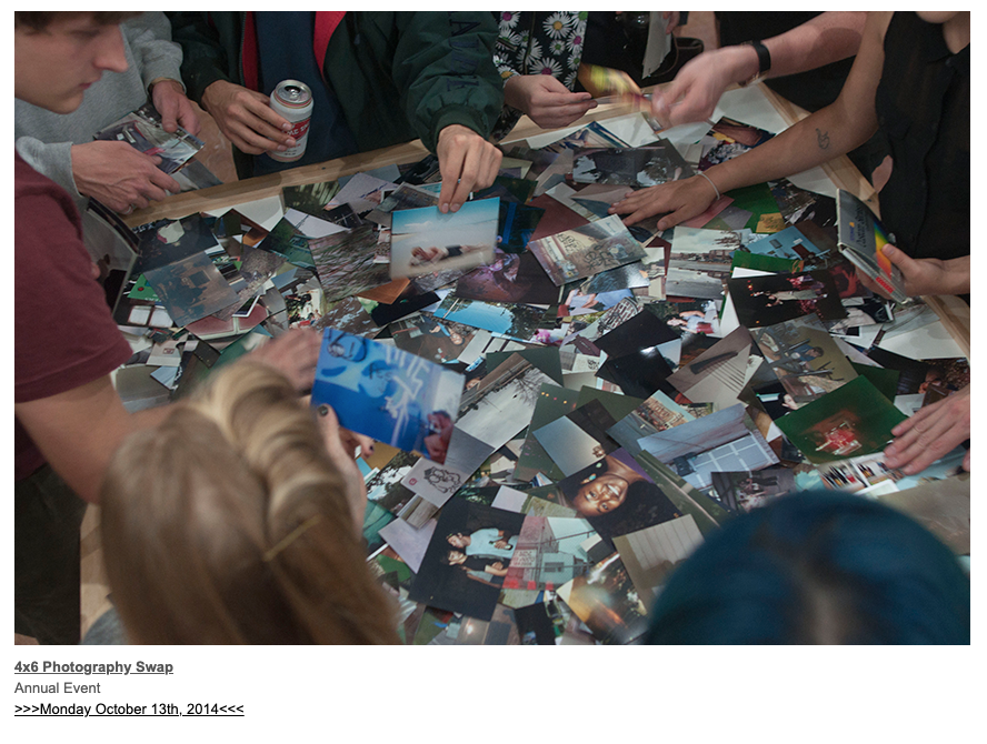 A photograph of several photographs scattered on a table. People surround the table, and several individuals hands' are reaching towards certain photographs.