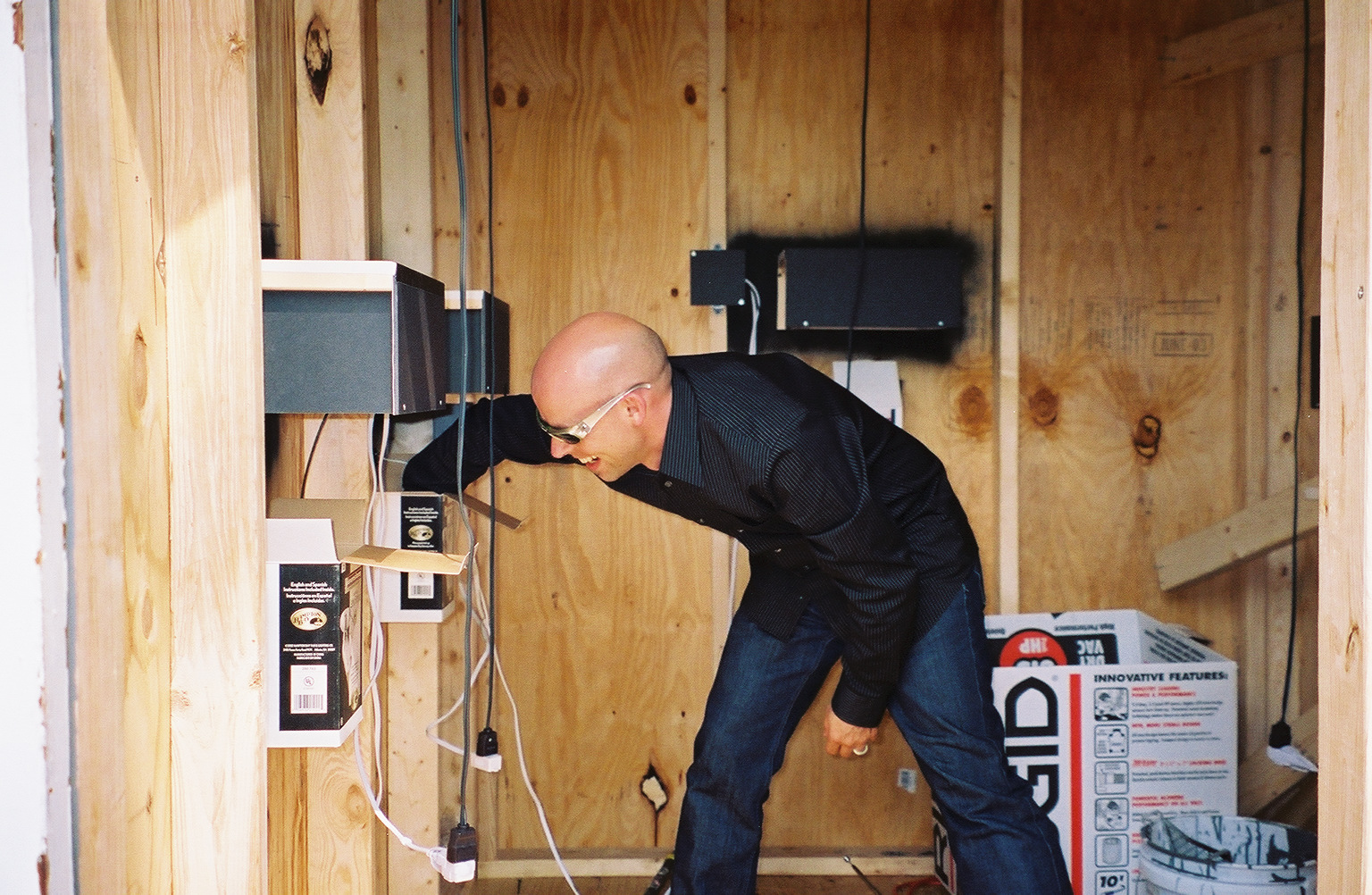 A photograph of a smiling individual in a black shirt and dark jeans reaching into a box against the wall, which is comprised of light-colored unstained wood.