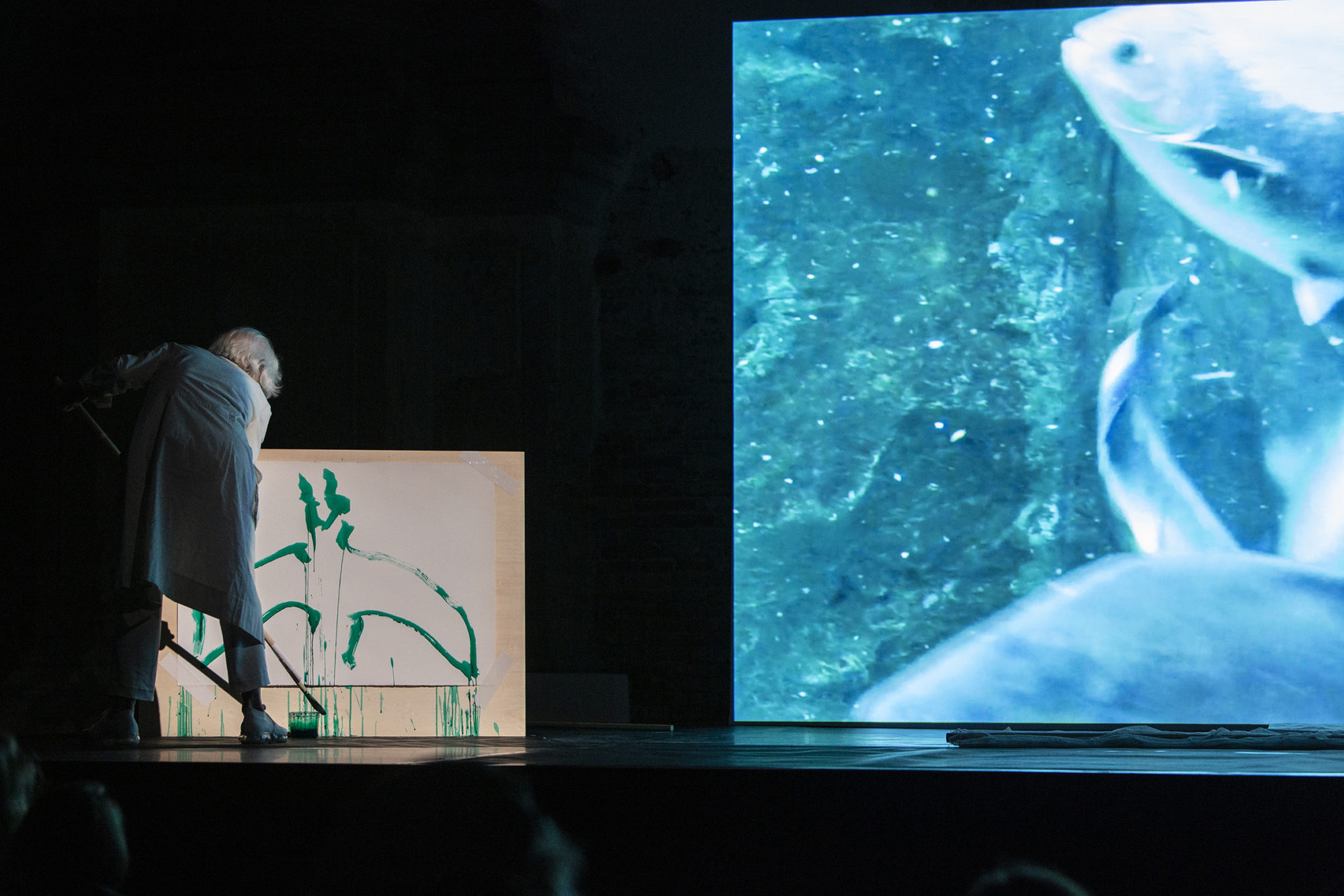Joan Jonas, an older white woman artist, performing in front of an aquarium projection and a green painted fish.