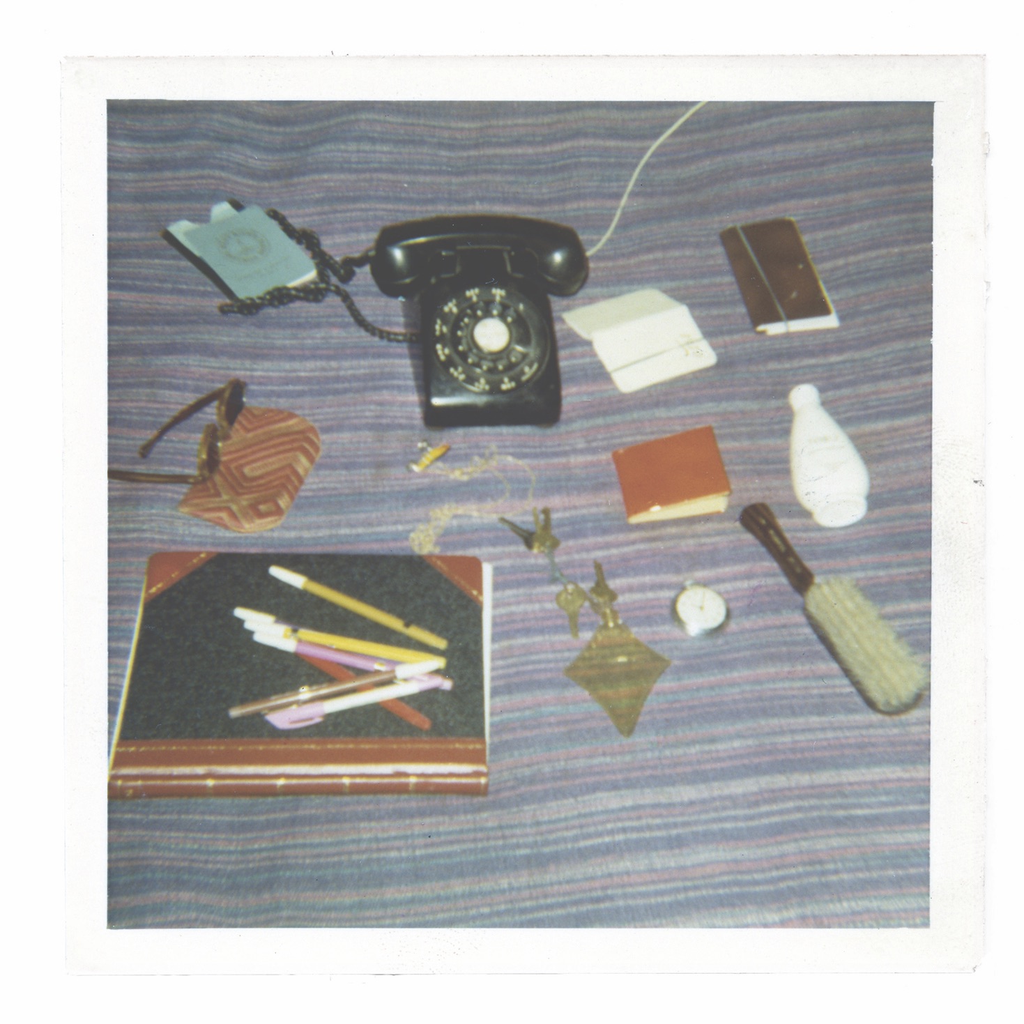 A photograph of several objects laid on top of a horizontally striped fabric. Objects include a dial phone, sunglasses, a hairbrush, keys, a notebook, pencils, a passport and a pocket watch.