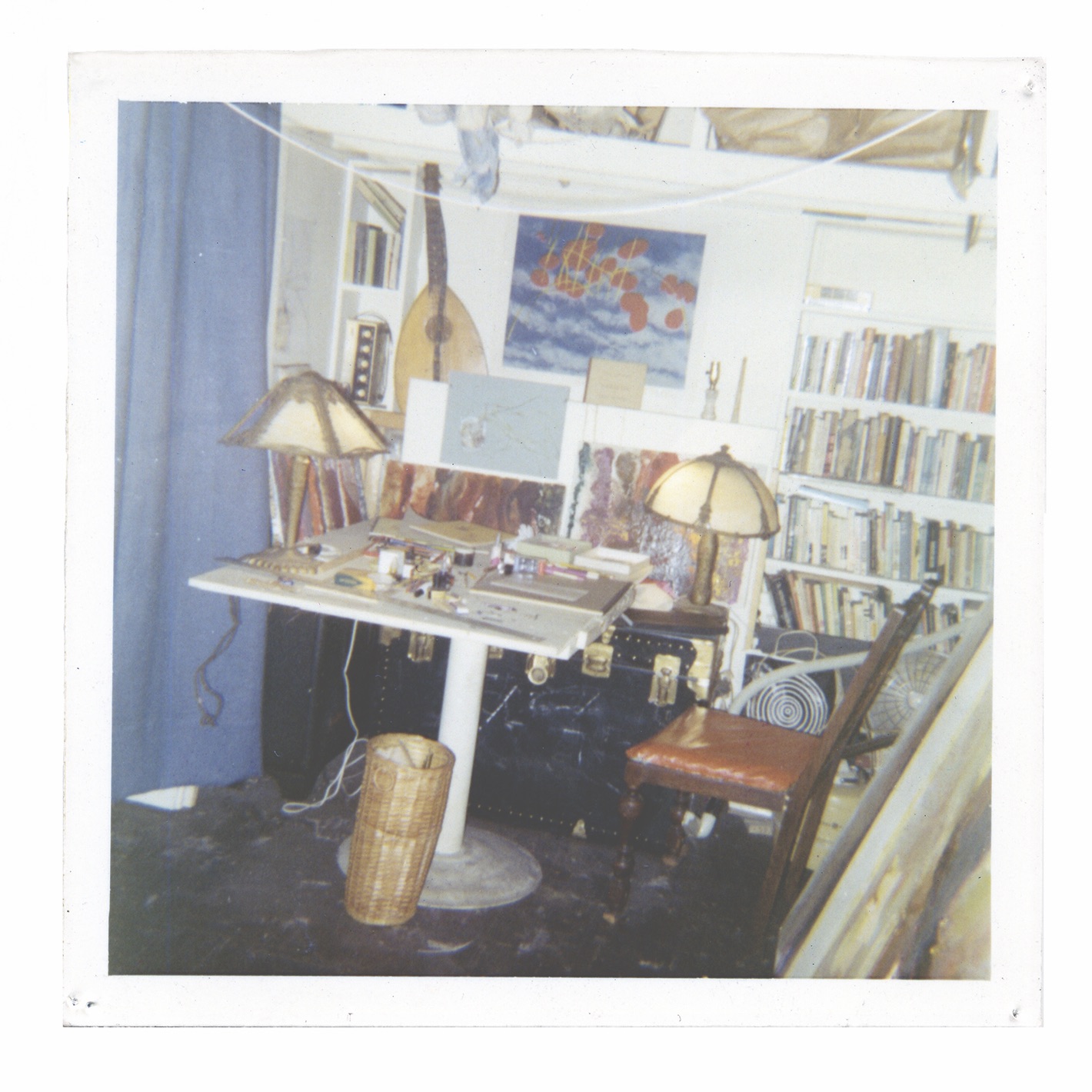 A photograph of a workspace, depicting a cluttered white table, a wood and leather chair, two cream-colored lamps, a bookshelf, a mandolin-like instrument and a blue curtain.
