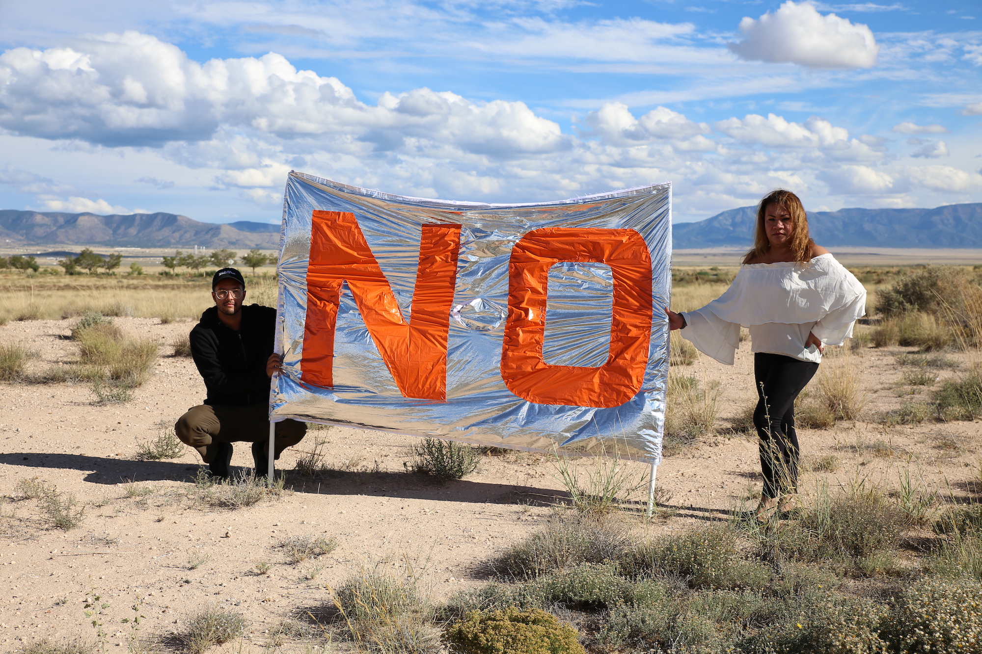 two people stand in the desert with a banner saying 