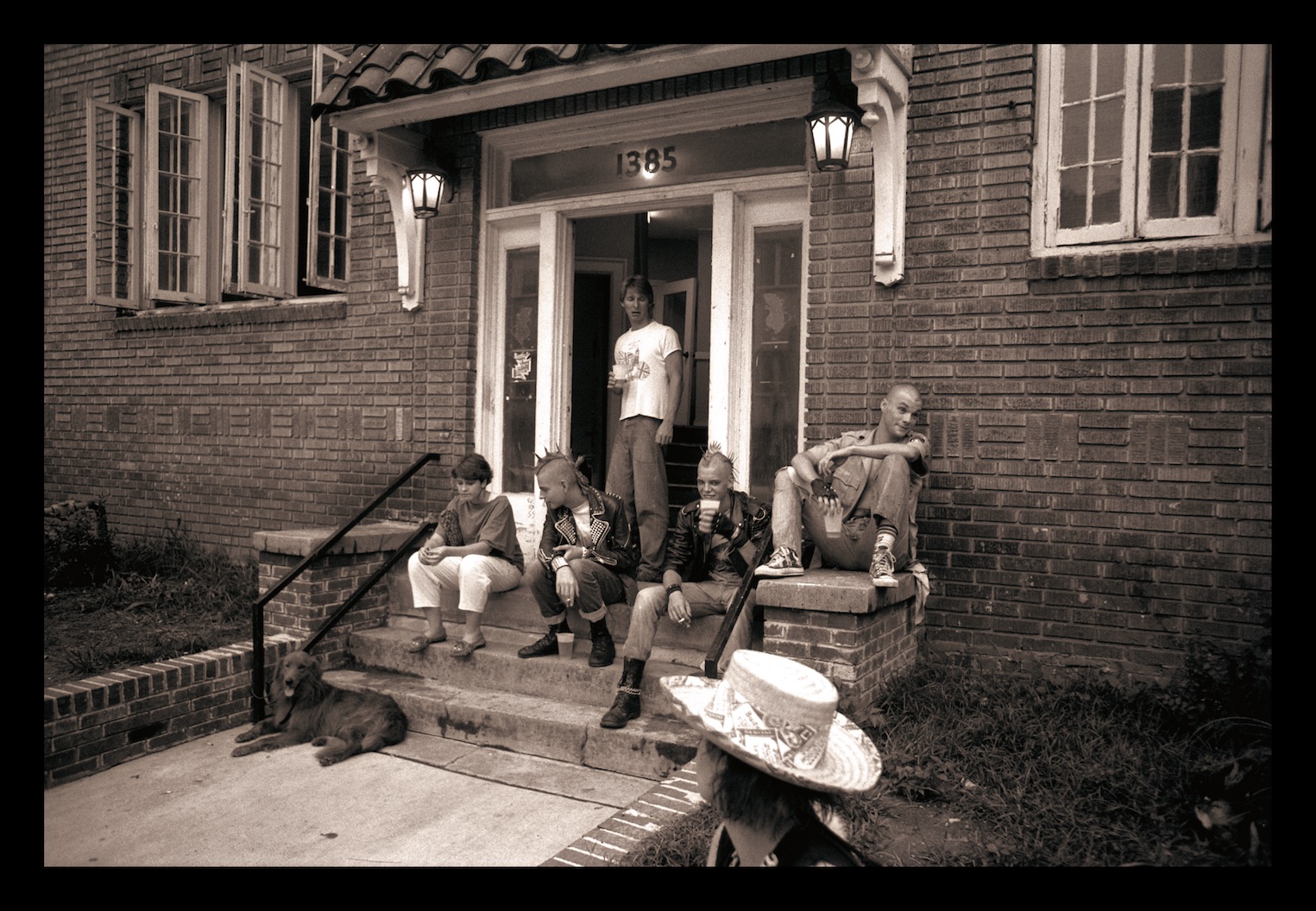 A black and white photo of four individuals sitting on the steps outside a brick building.