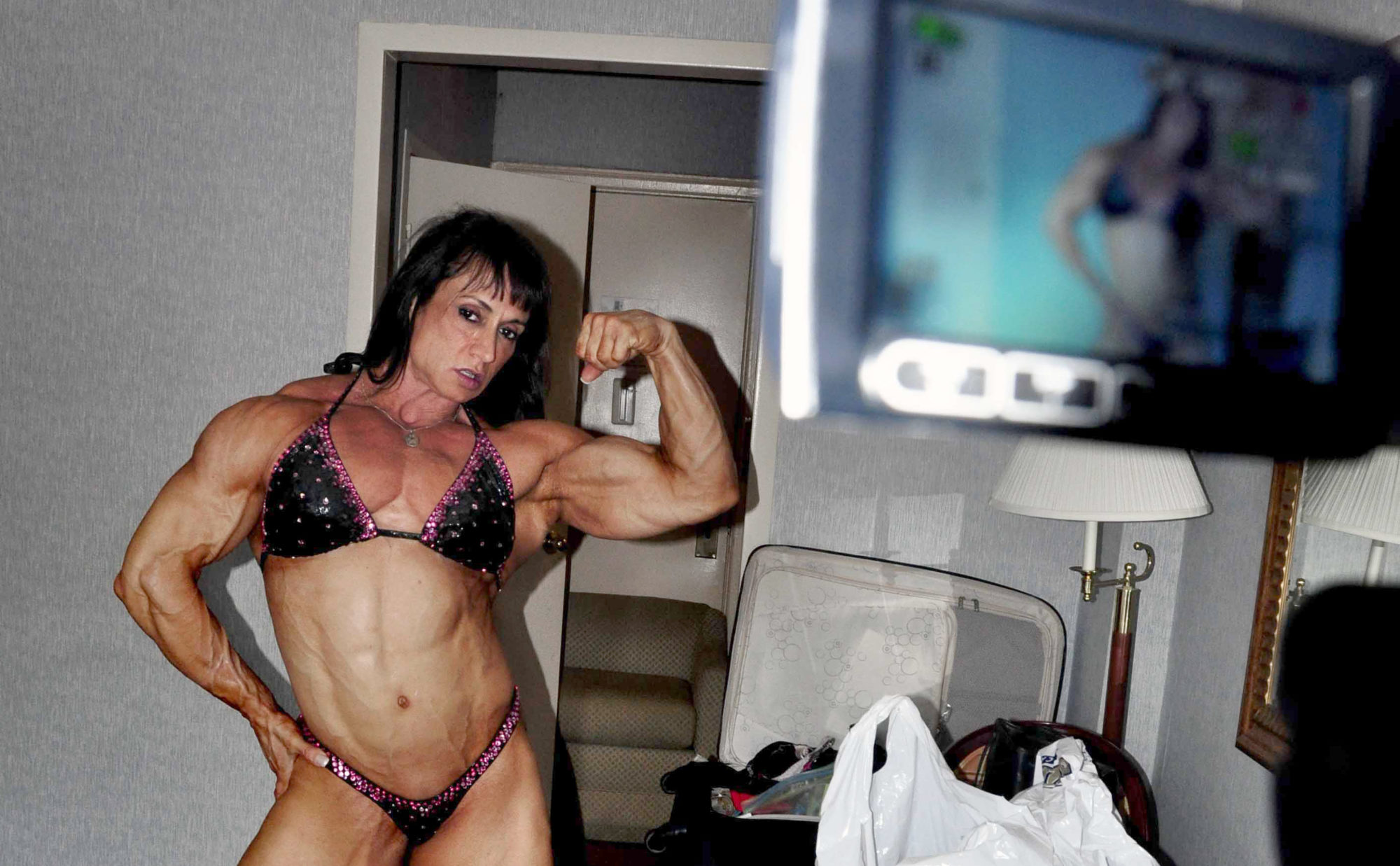 bodybuilder in bikini flexes bicep in front of video camera in a hotel room