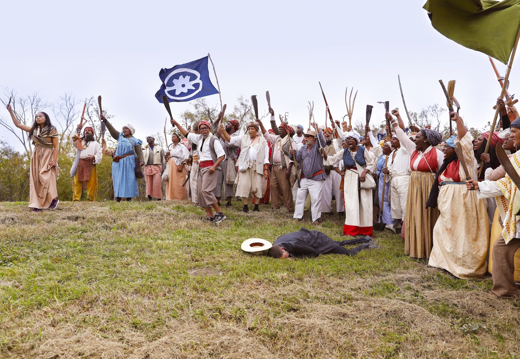 A White man lays (mock) dead in front of a group of Black men and women in historical dress. They raise their guns, machetes and pitchforks in triumph as they perform a slave rebellion reenactment