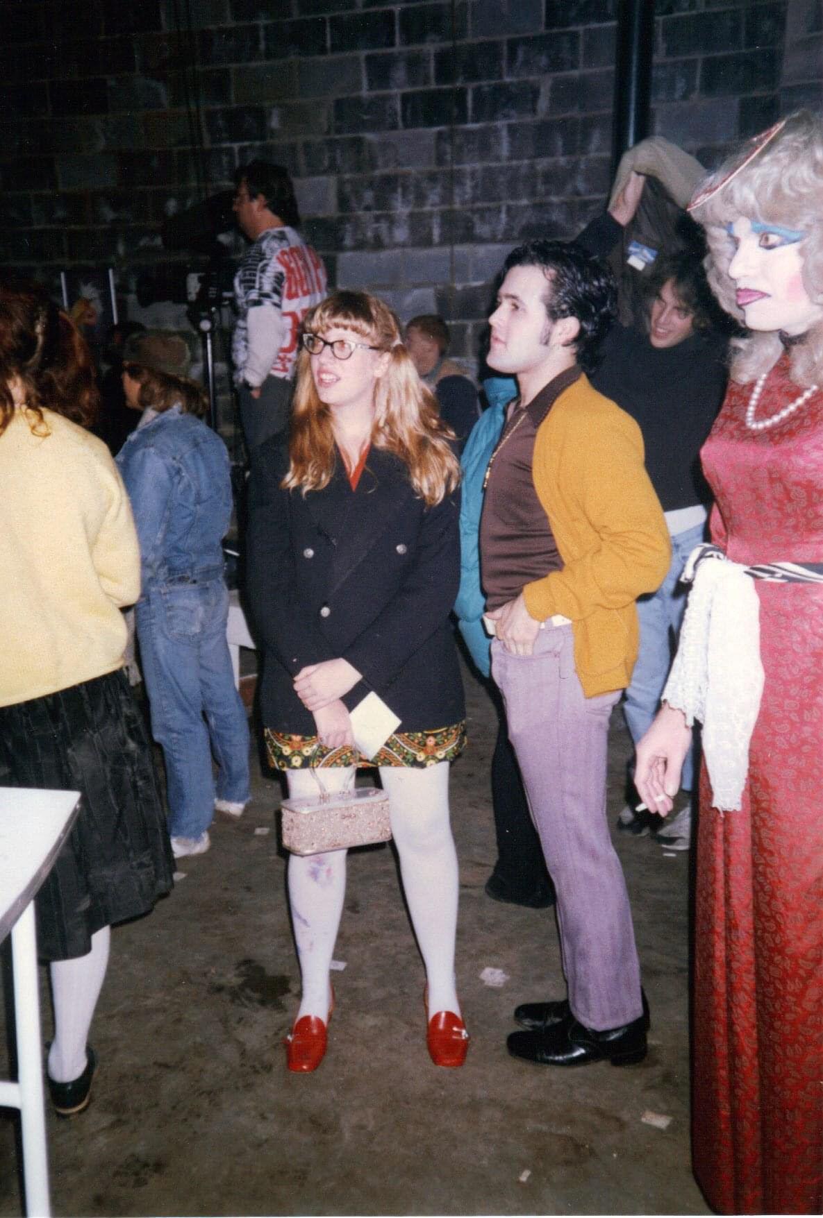 A photograph of several individuals in a dark space. One is dressed in a long red dress, a pearl necklace and a wig. Another is dressed in a dark glazer, white stockings and red shoes.