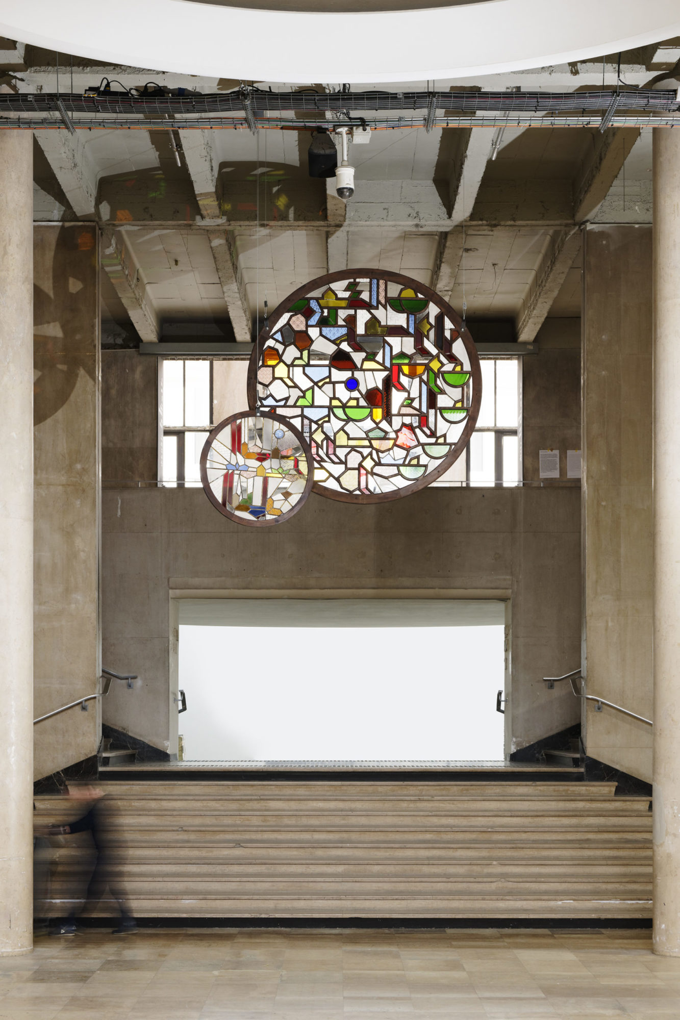 two circular stained glass windows by Sara Ouhaddou hang in an open beige concrete stairwell; the smaller window hangs to the left and slightly in front of the larger one