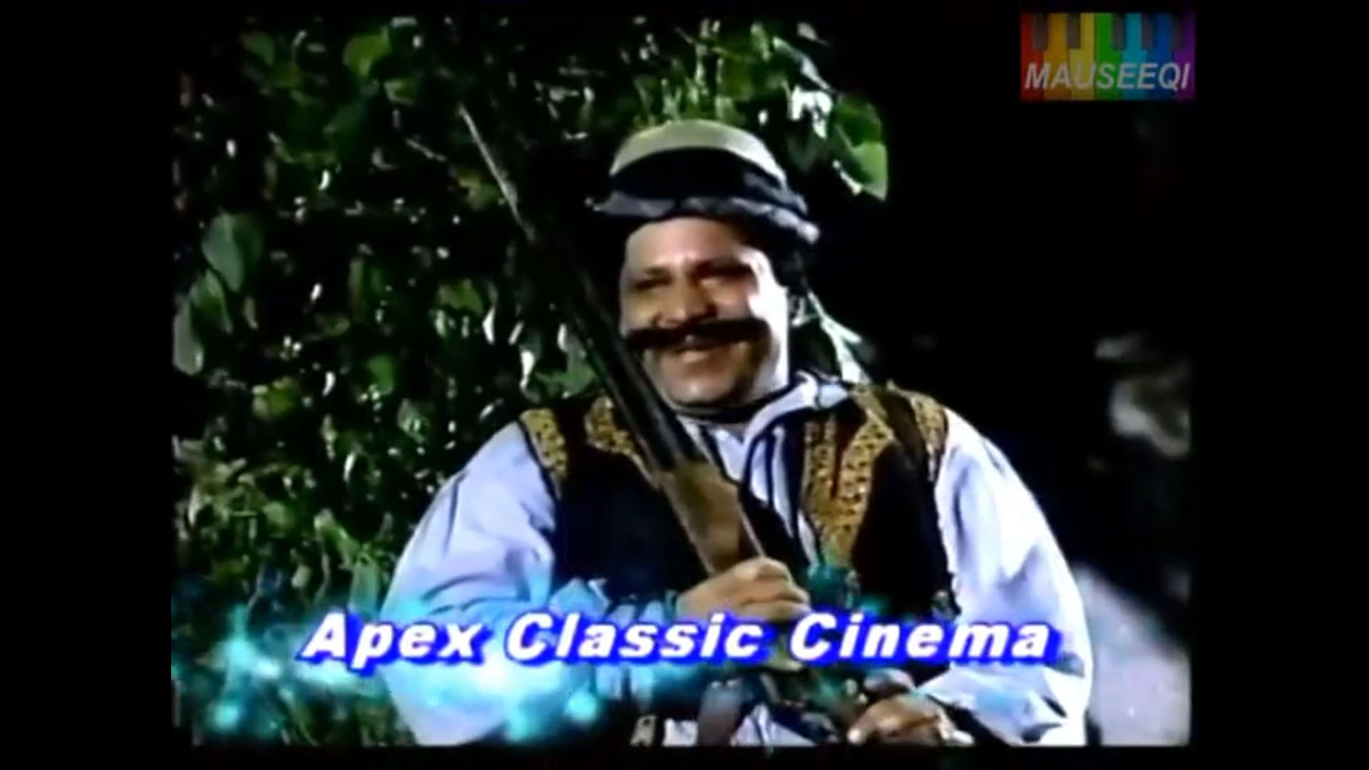 A screenshot from Dr. Bukhari's archives; A Pakistani actor is holding a rifle against his shoulder and smiling; the logo in the top right reads 'mauseeqi' and the banner across the bottom reads 'Apex Classic Cinema'