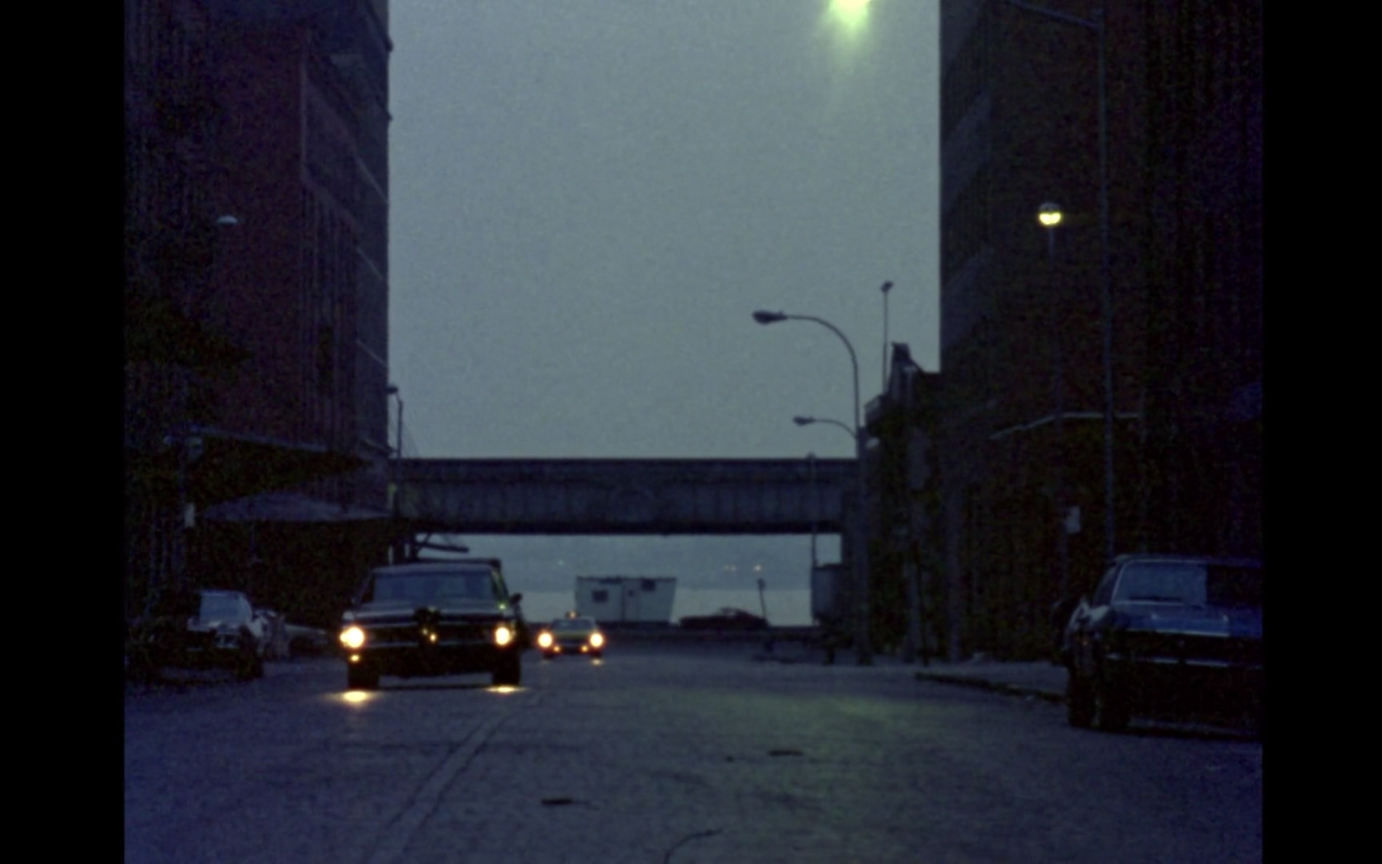 Screenshot from film News From Home displaying buildings, a bridge, and a car driving forward.