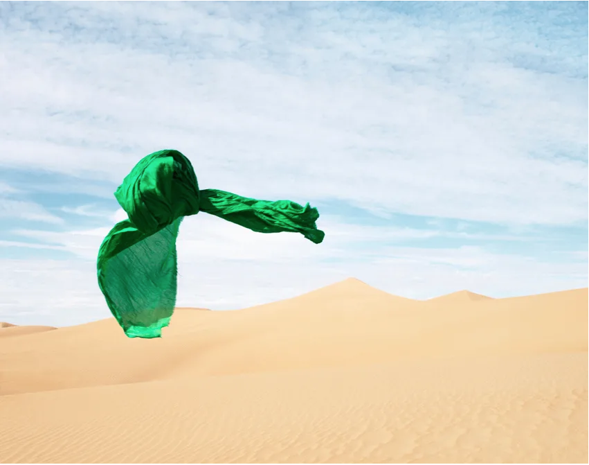 A vibrant green cloth is suspended above rolling sand dunes set against a blue and partially cloudy sky