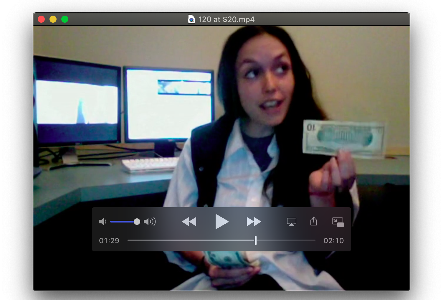 A screenshot of a video in which a person with long hair and a white shirt is holding a ten dollar bill upside down. The bill is pressed between her index finger and thumb, and the person is gazing up and to the left.