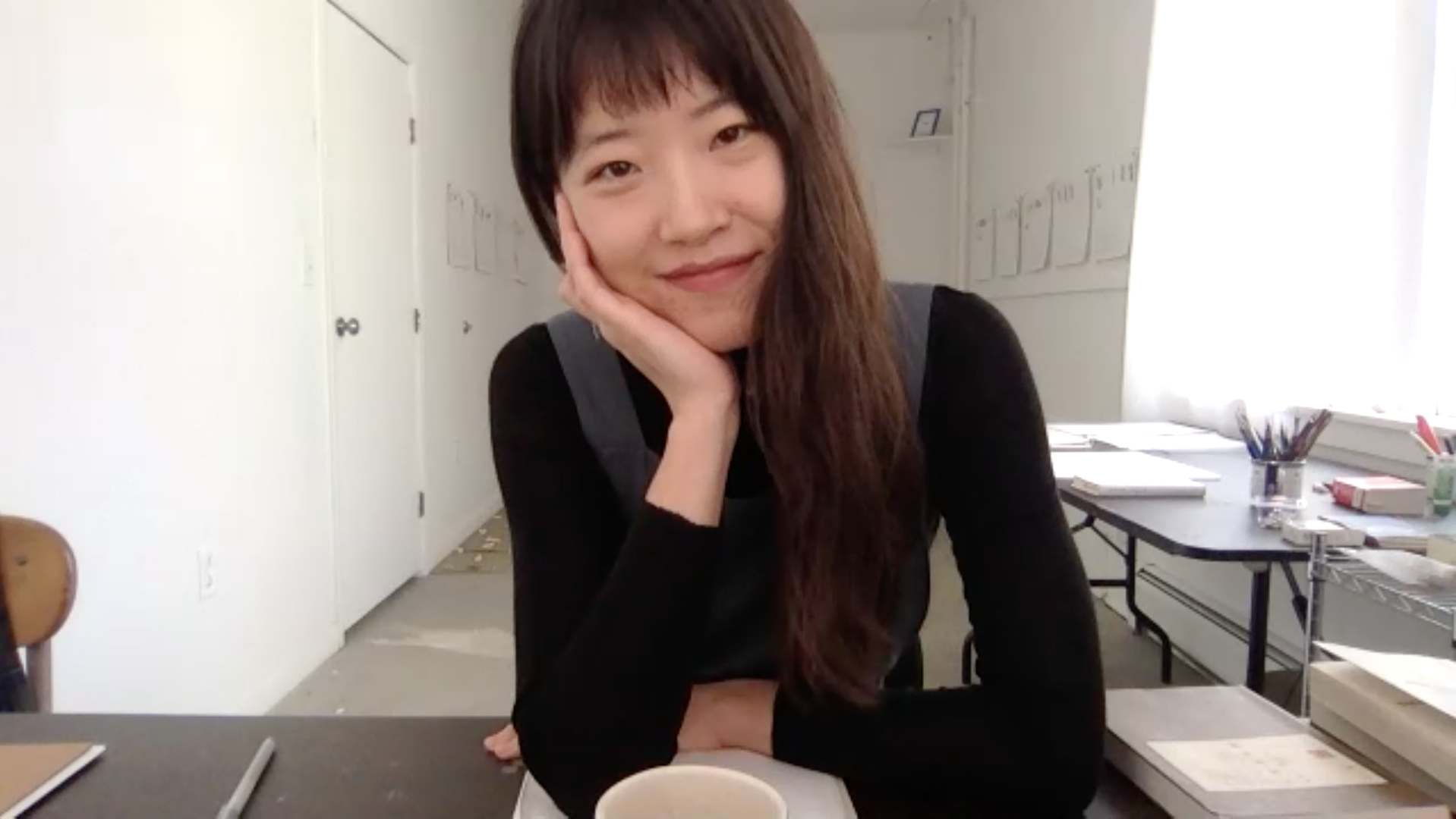 Screenshot of Chang Yuchen sitting in her studio at her desk during the interview, with her left arm resting on the desk and her head resting in her right hand, smiling into the camera.
