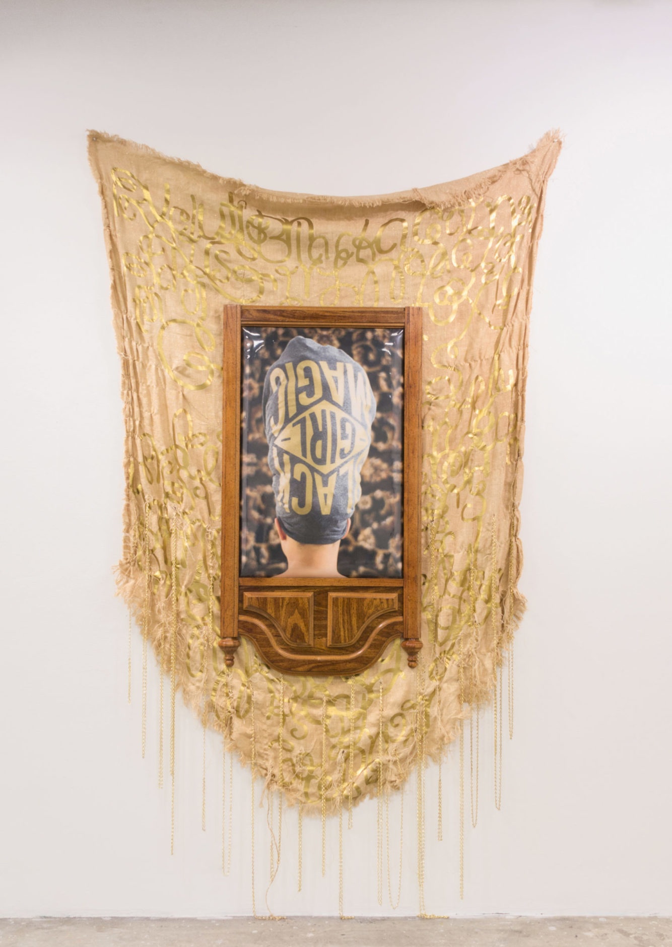 An asymmetrical gold-painted fabric hangs behind a wooden dresser mirror frame; inside the frame is a photo of the back of a black woman's head wrapped in cloth that reads 