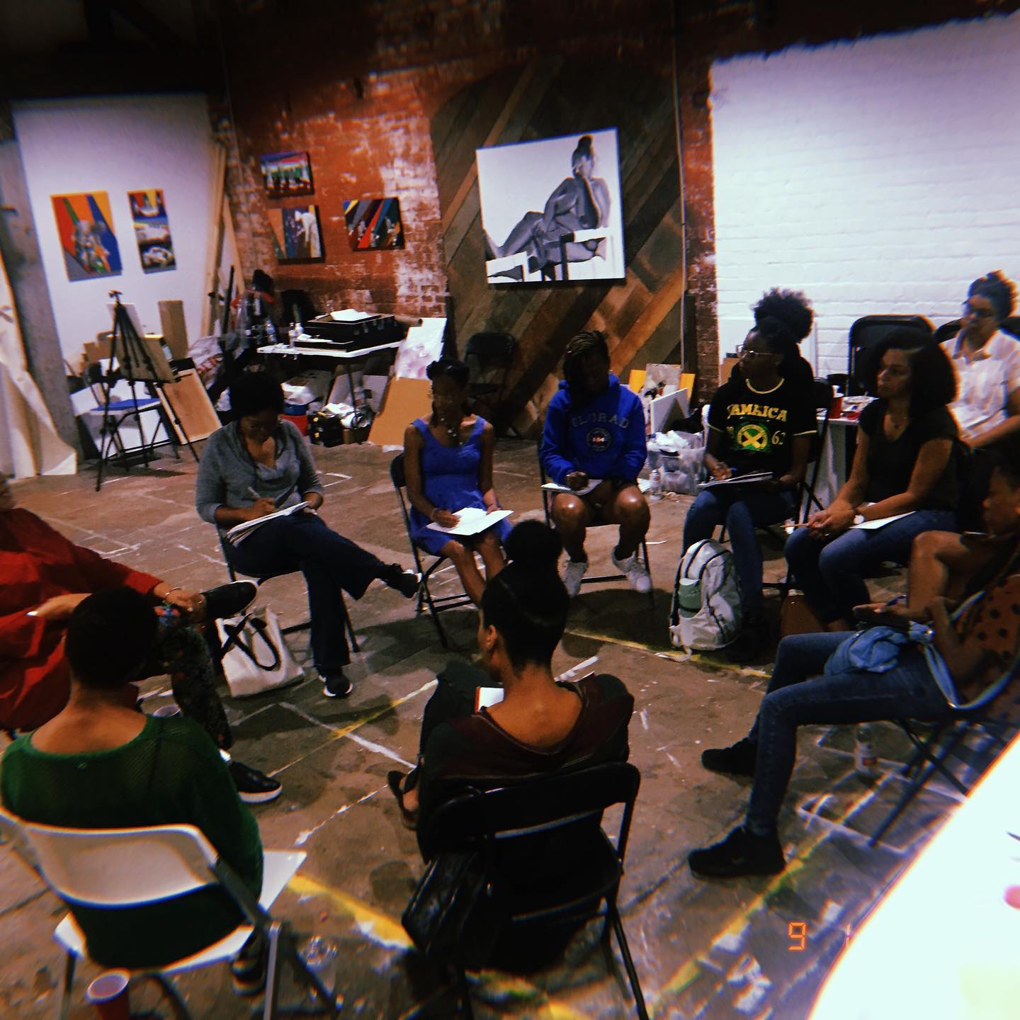 A photograph of around ten individuals sitting on folding chairs in a circle with notepads in their laps. There is art on the walls and a concrete floor.
