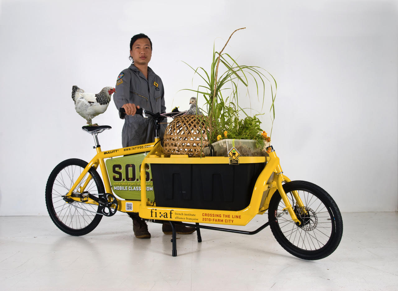 Tattfoo Tan stands next to a yellow bicycle cart which holds a chicken, a basket, and plants