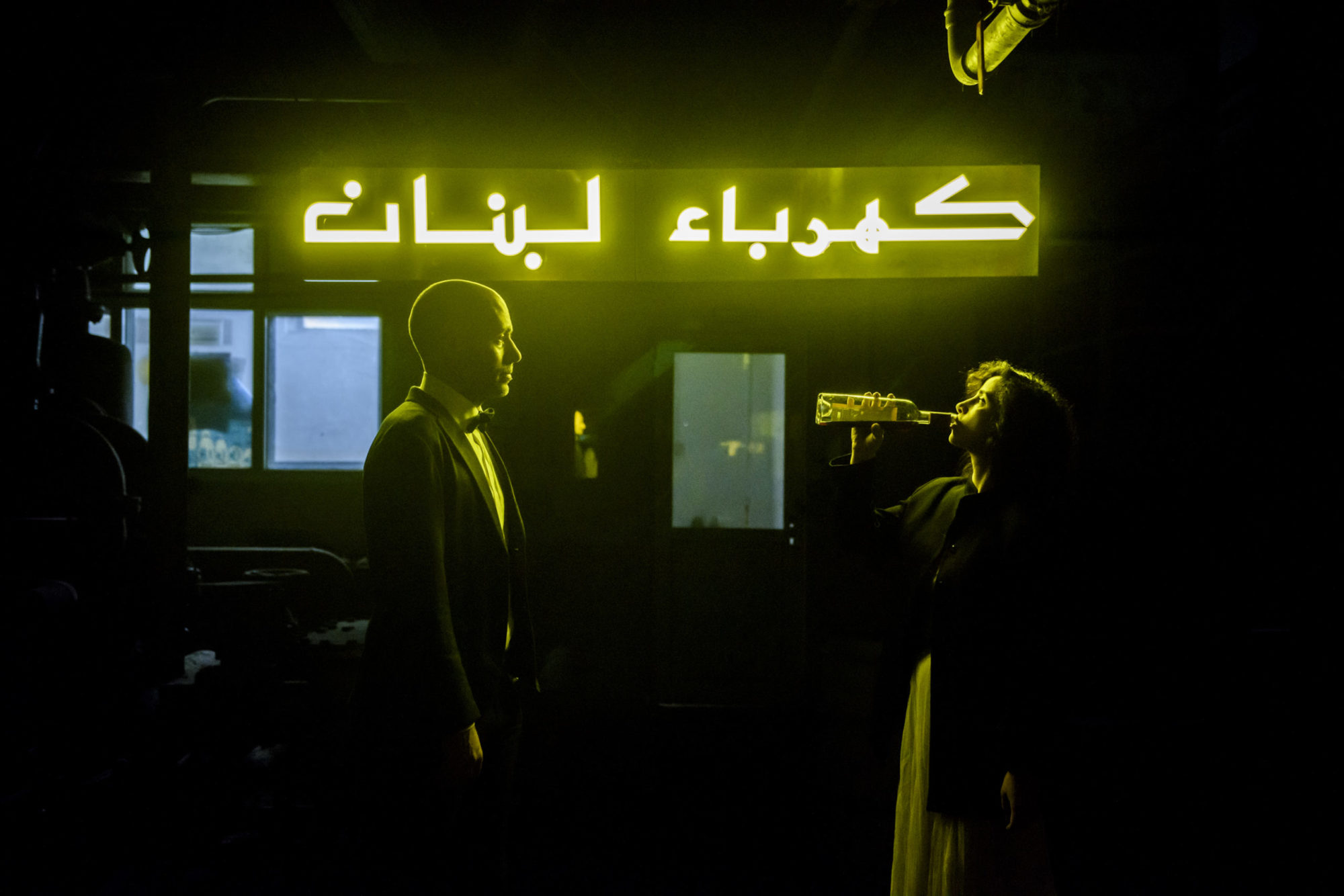a man and a woman face each other, back-lit by the green glow of a neon sign; the man, dressed in a tuxedo, watches as the woman opposite him takes a swig straight from a bottle