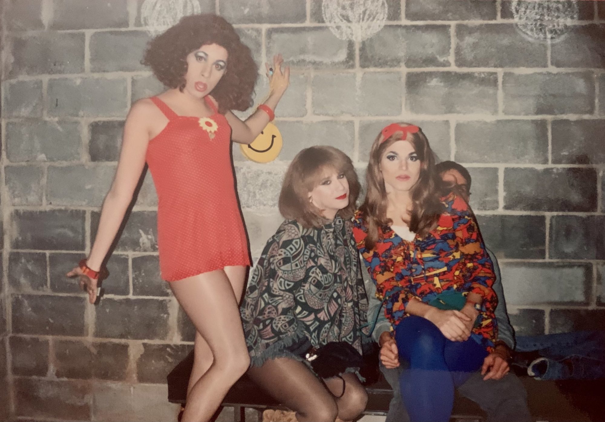 A photo of three individuals dressed colorfully — bold, metallic makeup and bright red clothing —against a gray brick wall.