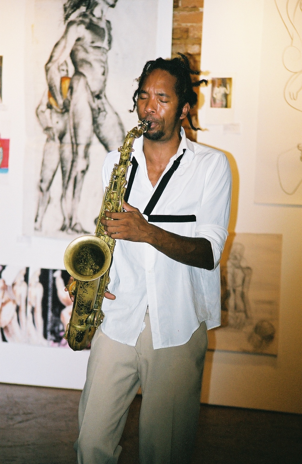 A photo of an individual wearing a white shirt and khaki pants playing the saxophone in Apache Cafe — several works of art hang on the wall.