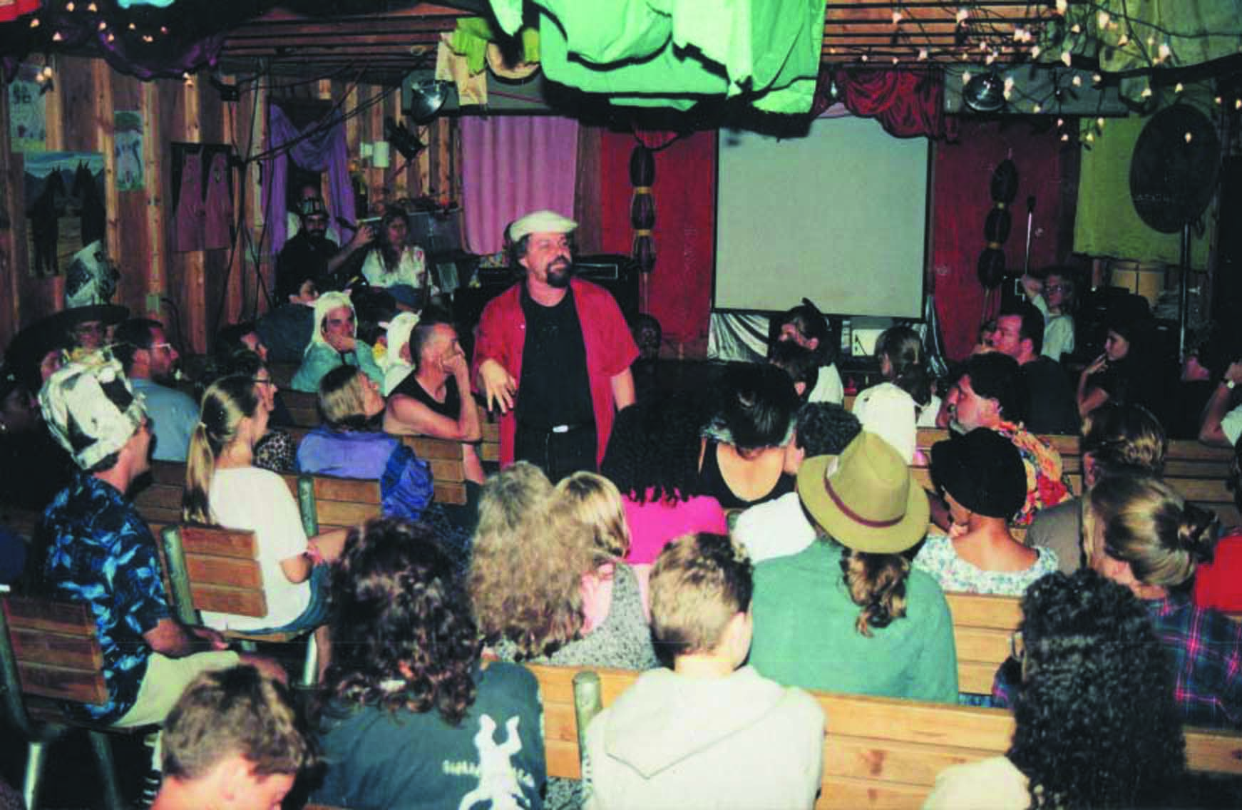 A photo of an event at Cafe Bizzozos. The audience is seated on wooden benches, and colorful art hangs from the ceiling and walls.