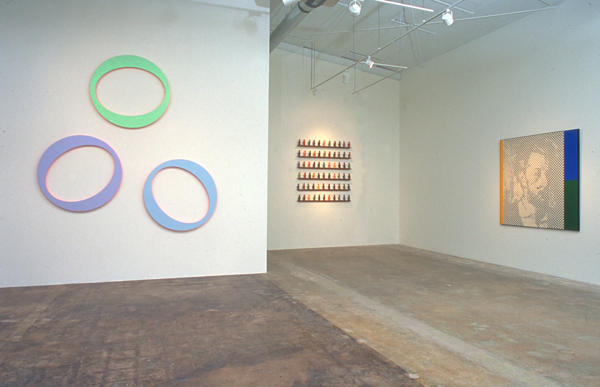 A photograph of an installation view. On the left are three round pieces of art, colored green, purple and blue. On the right is a pixellated rendition of a face flanked by blocks of yellow, blue and green.