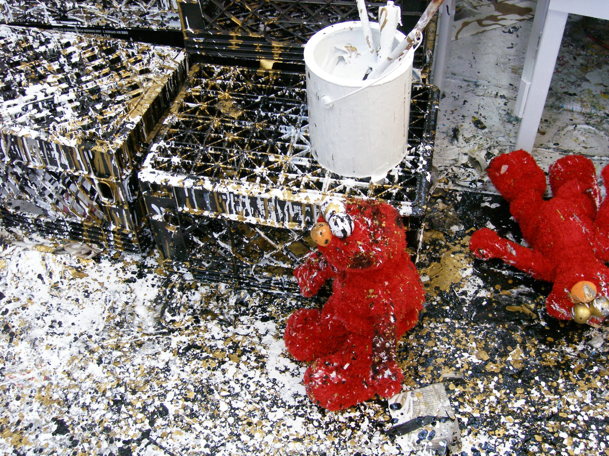 Photo contains stacked black crates, a pure white paint can and elmo dolls, all covered in a thick splatter of white, gold and black paint.