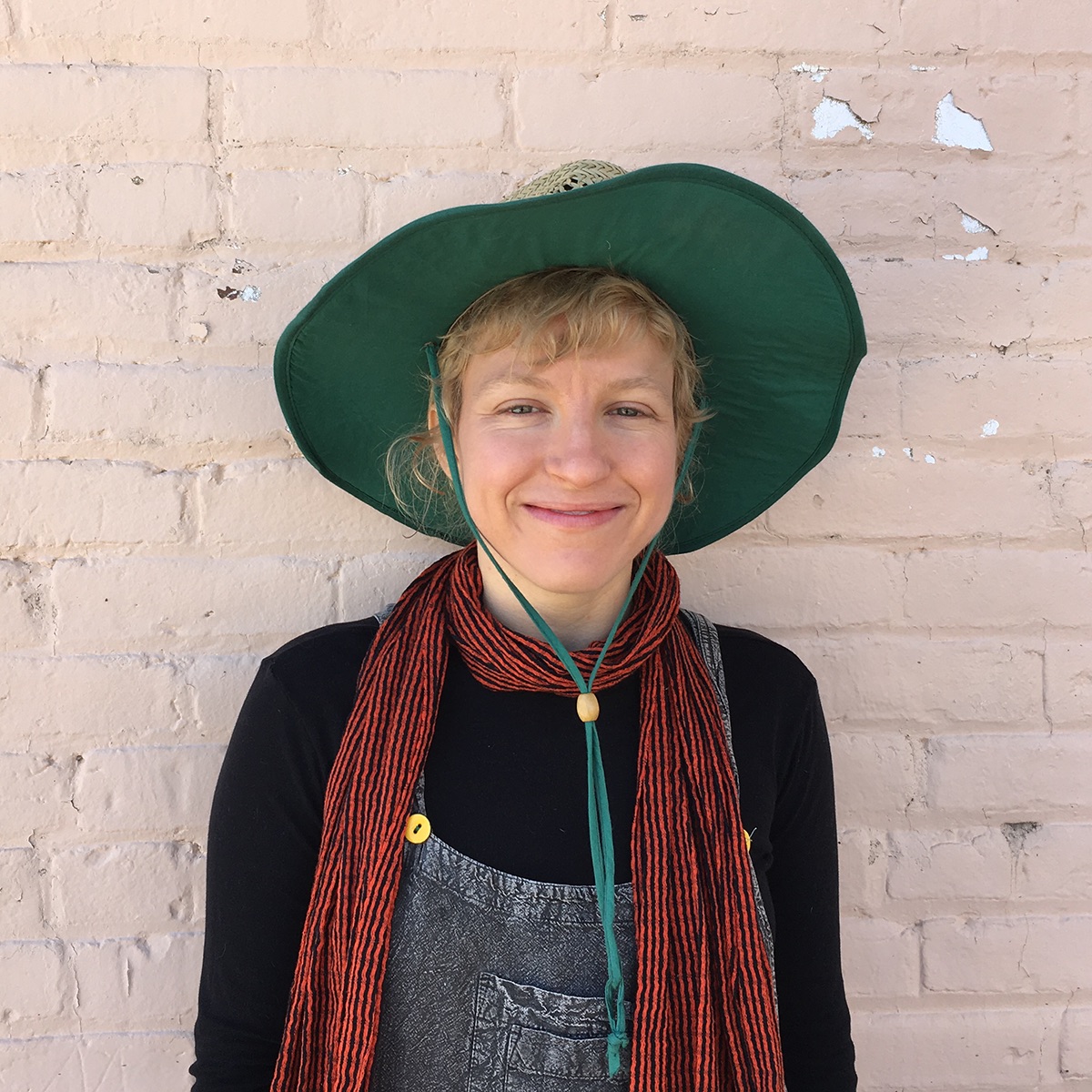 A portrait shot of a white nonbinary person with short blonde hair in their thirties, standing and smiling against a beige brick wall, wearing a hat with a big green rim, overalls, and a black and orange scarf.