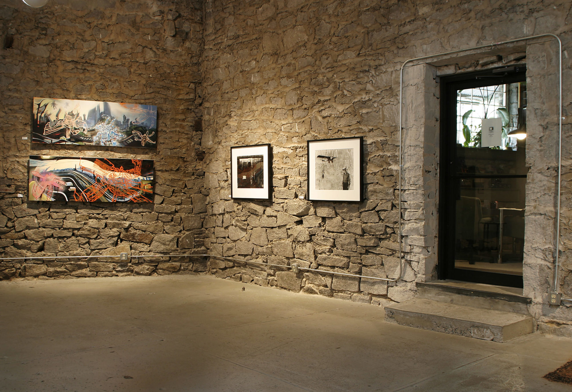 A photo of an installation, with a concrete floor and stone walls. Four pieces of art hang on the wall, and there is a door on the right side of the image.