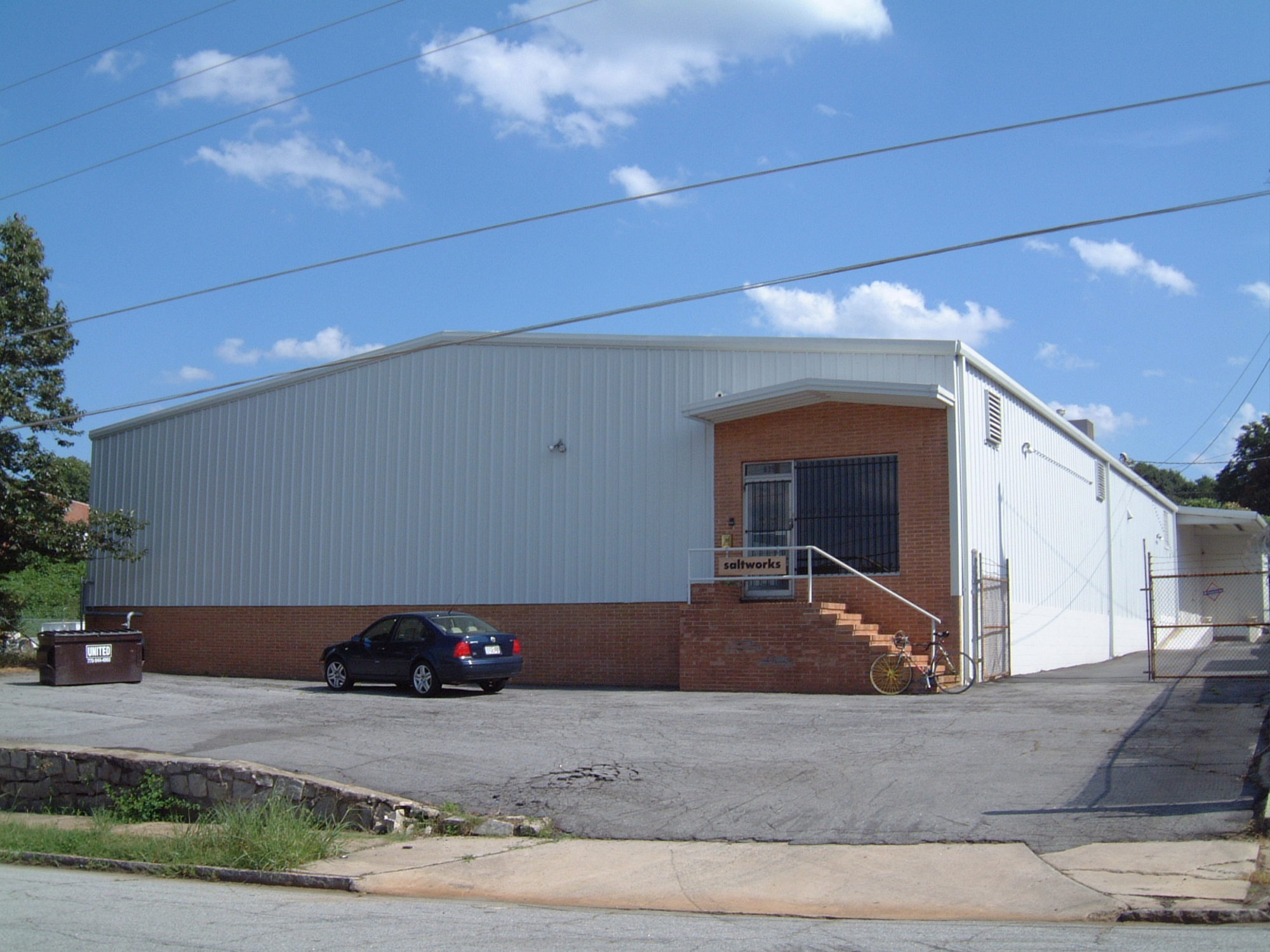A photo of the exterior of the Saltworks building. A few brick stairs lead to a screen door, and the rest of the building is corrugated metal. It's a sunny day with a few fluffy clouds.