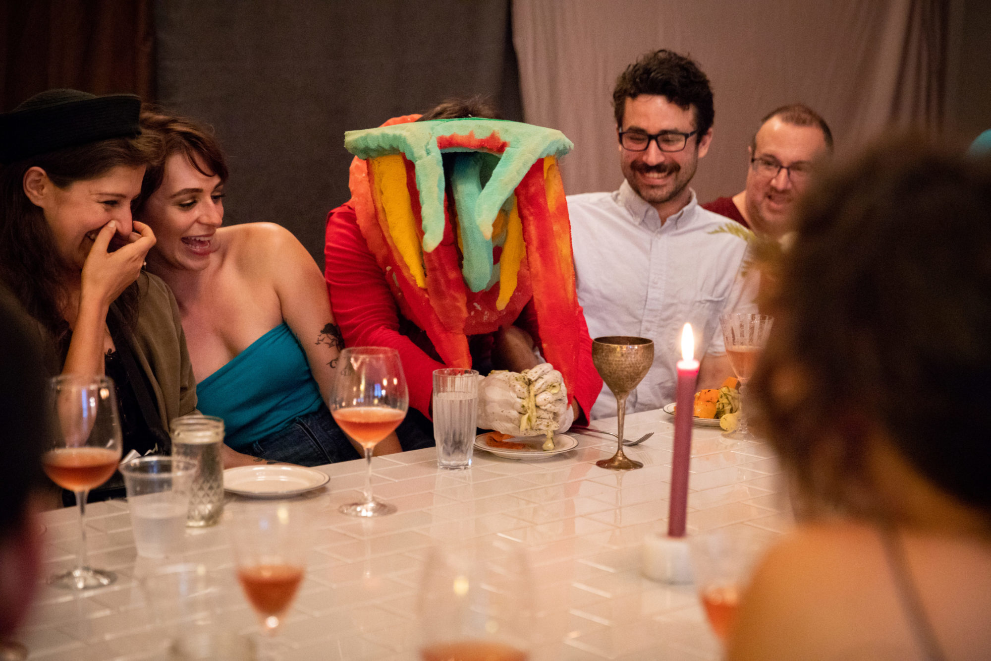 person in red clothing and wearing a mask plays with food at a table with other people