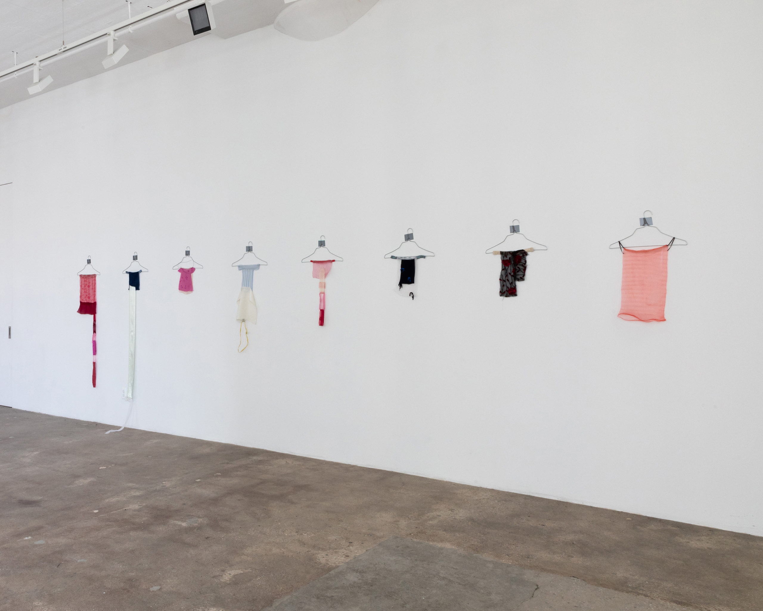 There are eight hangers on a plain white wall with tops and other pieces of fabric hanging on them.