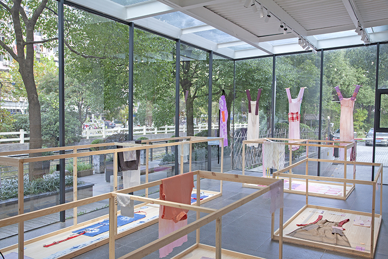 A room with floor-to-ceiling glass windows with a view of trees and greenery outside, and wooden fixtures inside that display garments and fabric. Four dresses are taped to the windows.