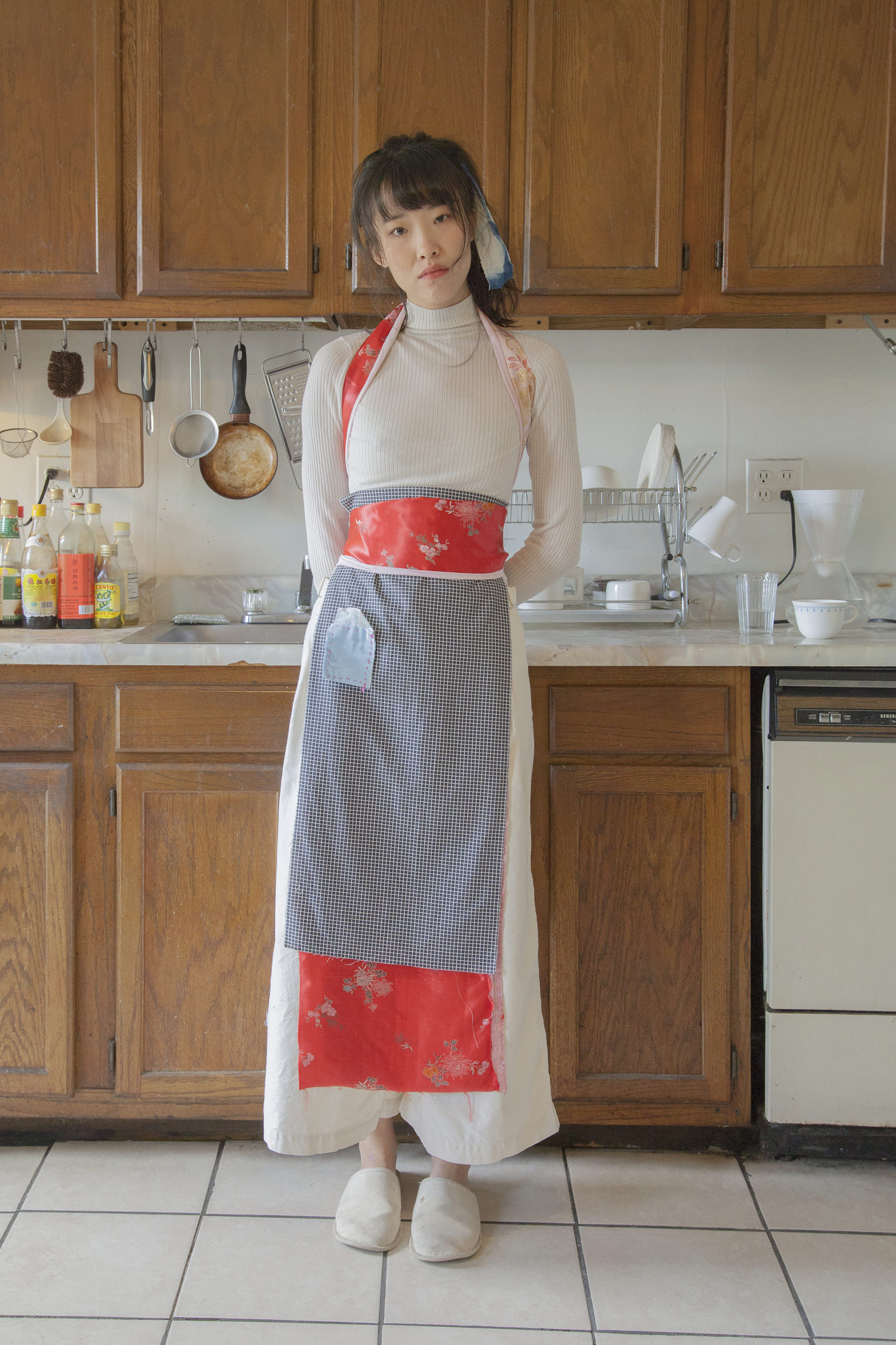 Chang Yuchen standing in a kitchen facing the camera wearing a beige top and pants with a peach-and-blue apron; the peach fabric is silky and the blue on the front part looks like cotton.