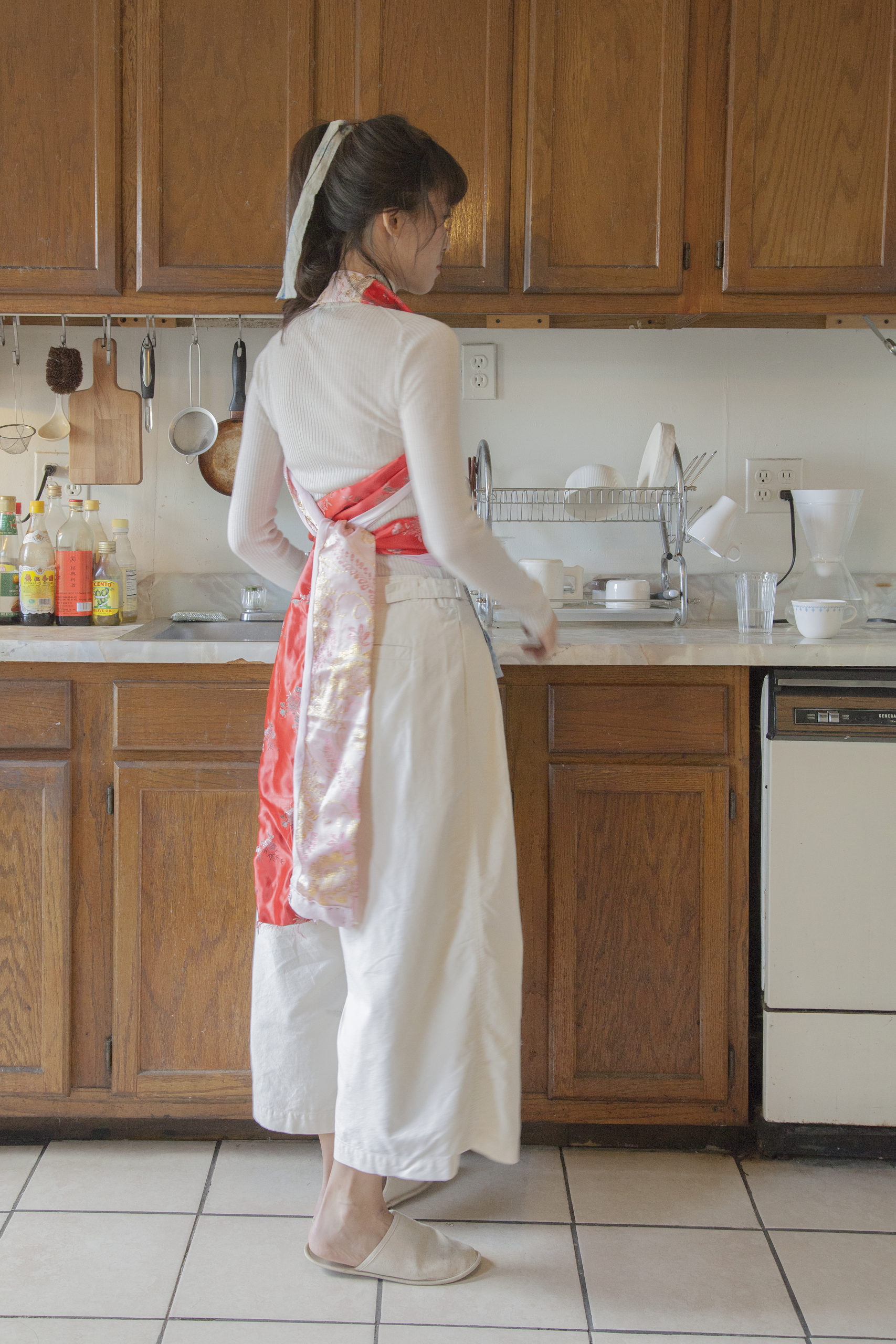 Chang Yuchen standing in a kitchen with her back to the camera, wearing a beige top and pants with a peach-colored apron that ties in the back.