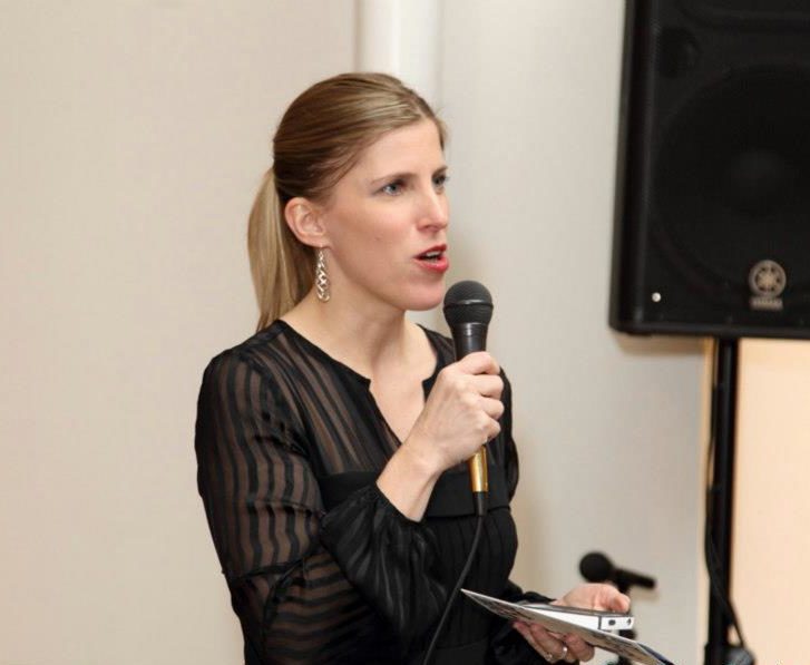 A blonde woman wearing black speaks into a microphone
