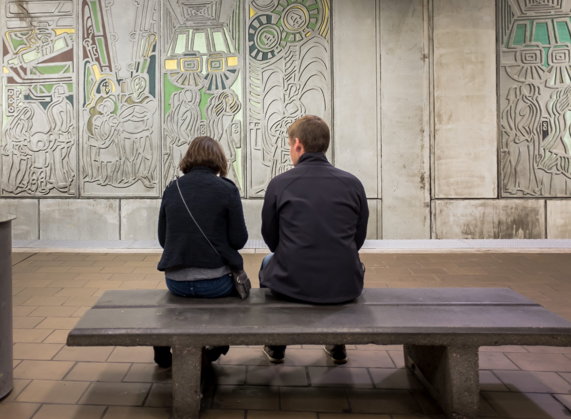 A man and woman sit on a bench facing a carved mural