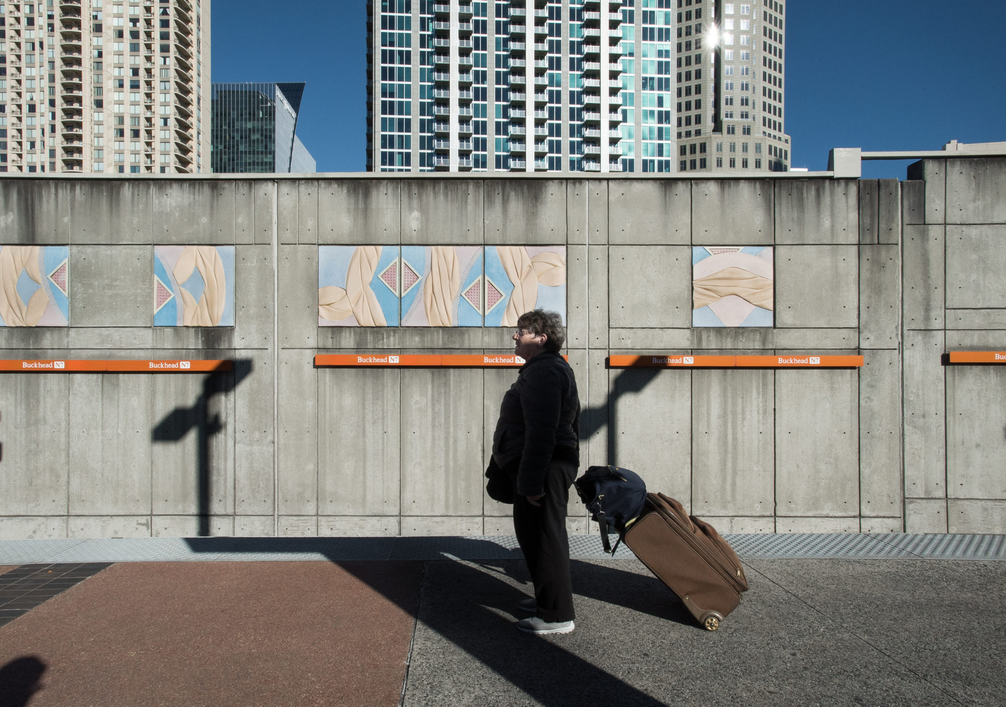 A person stands and holds luggage at a train station outside with apartment buildings in the back