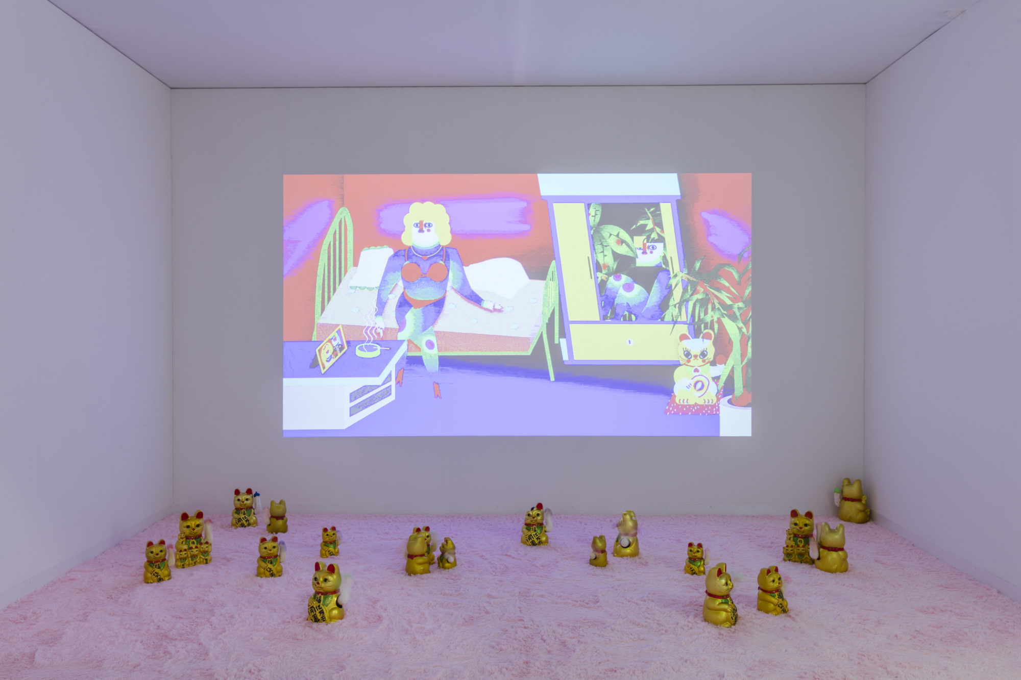 An installation with colorful artwork projected on the wall and fortune cats scattered on a pink carpet