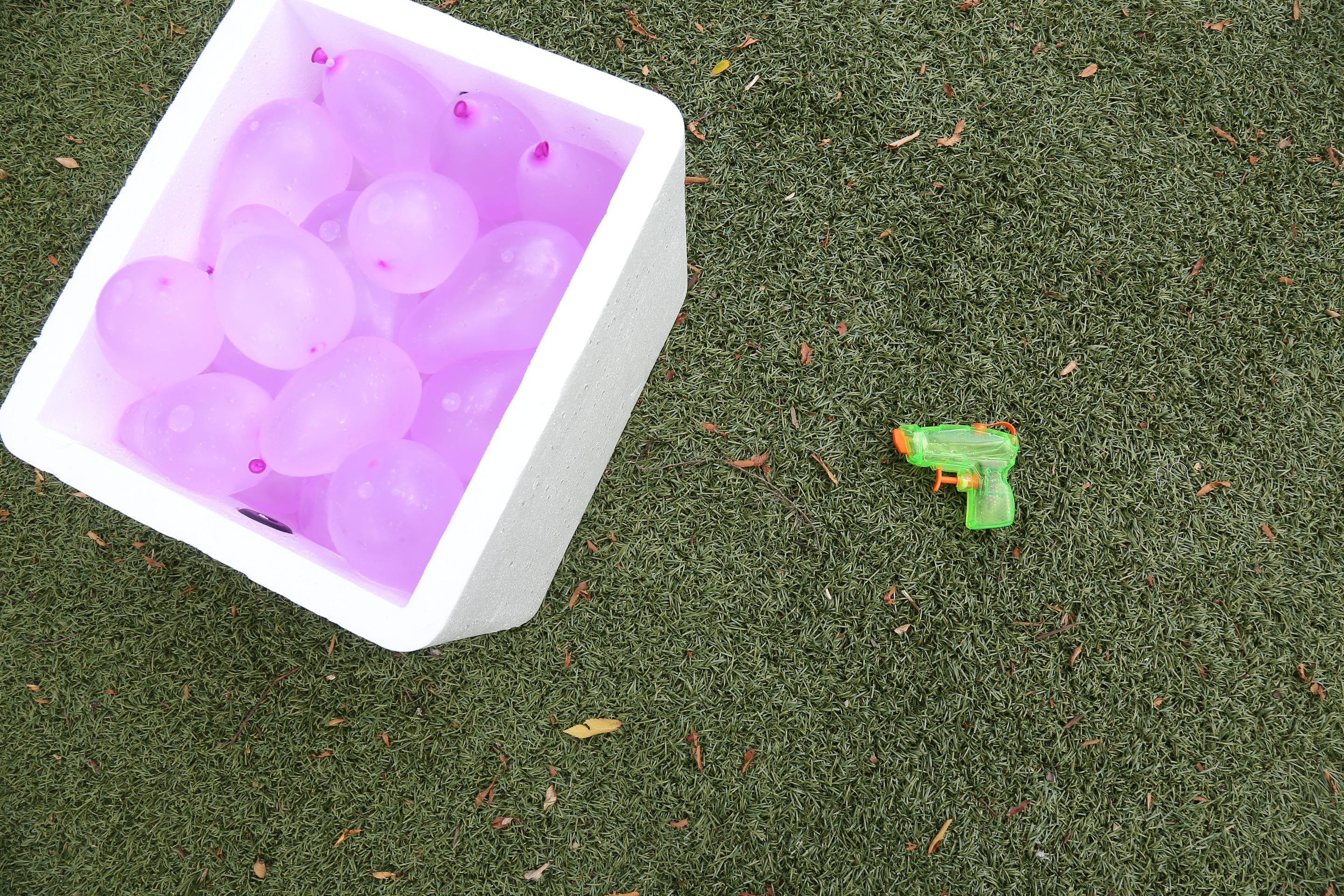 An image of a styrofoam cooler containing pink water balloons, sitting next to a tiny green water gun.