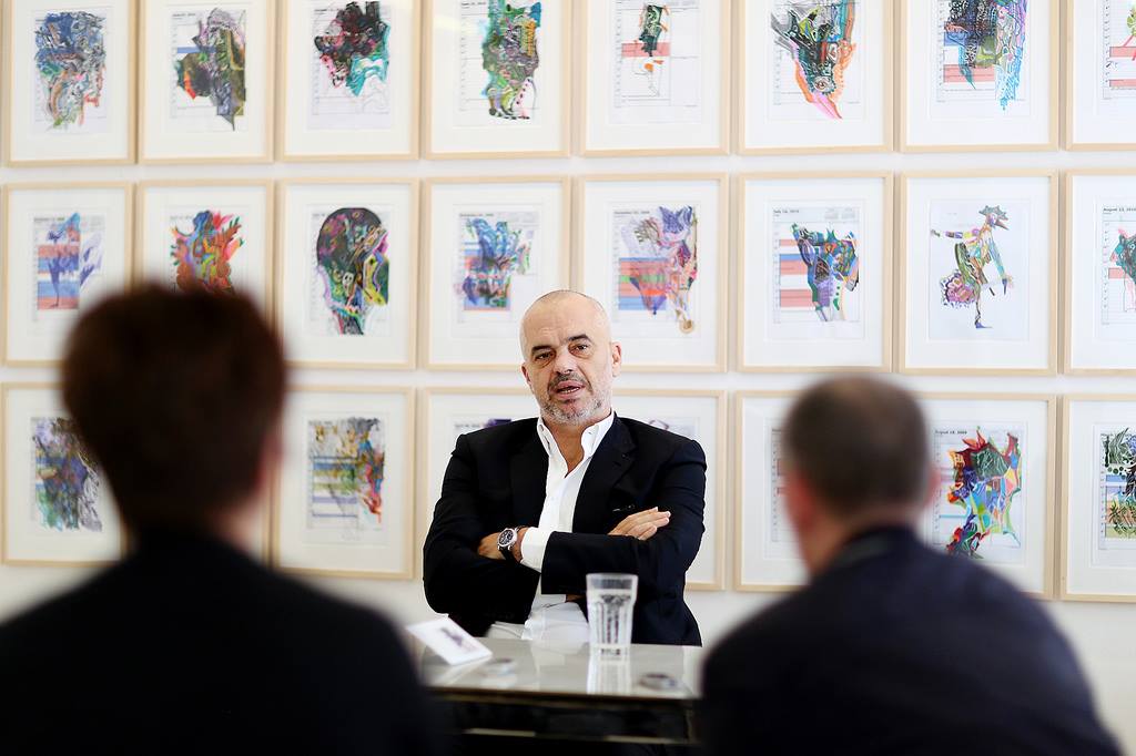 A man with his arms crossed sits in front of a wall of framed artworks and speaks to people