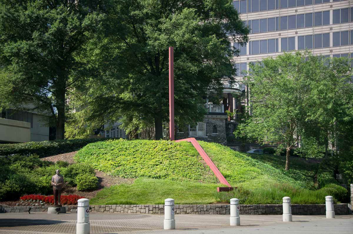 Long, red metal sculpture sitting on elevated grass hill in front of a building