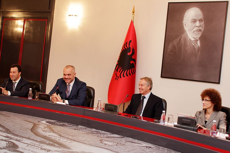 A woman and three men sit at a meeting table with a red flag and a framed portrait behind them