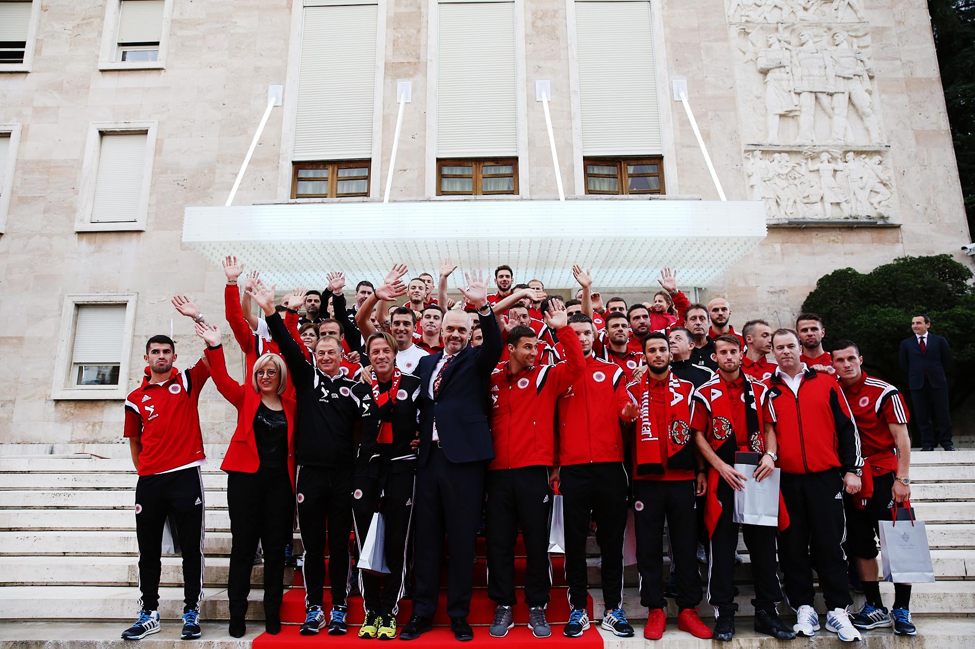 A man stands and waves alongside a soccer team clad in red clothing