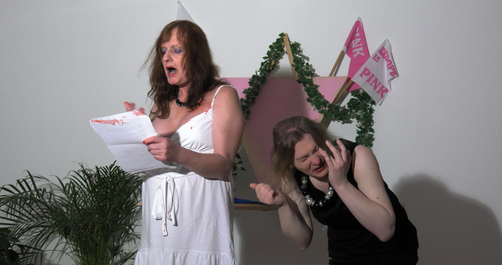 A woman in a white dress reads from paper while another woman in a black dress touches her face and grimaces