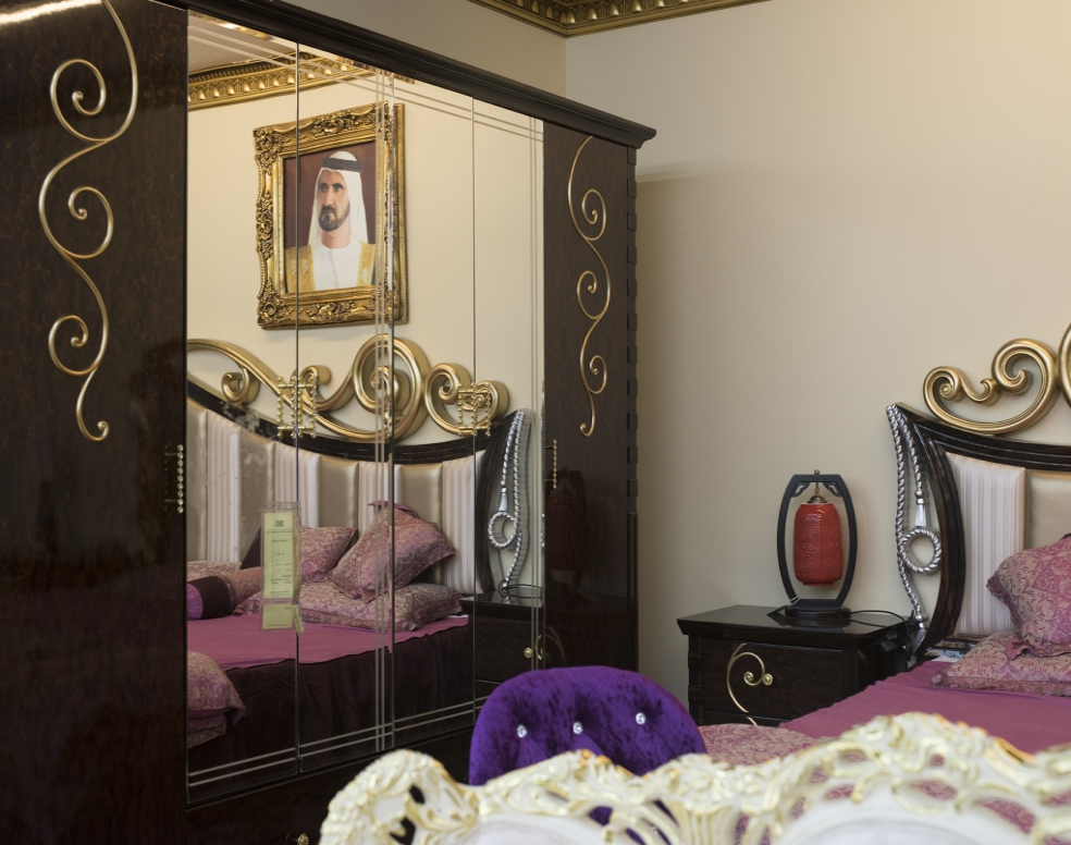 Dark brown wardrobe with gold swirls and mirror reflects matching bed and bedside table with a portrait of a man hanging on wall