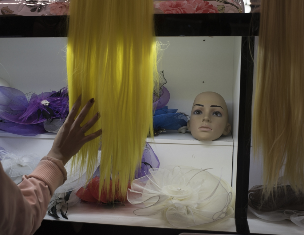 Hand brushes bright yellow wig hanging in front of black and white shelves with purple and blue decorative hats and a mannequin head.
